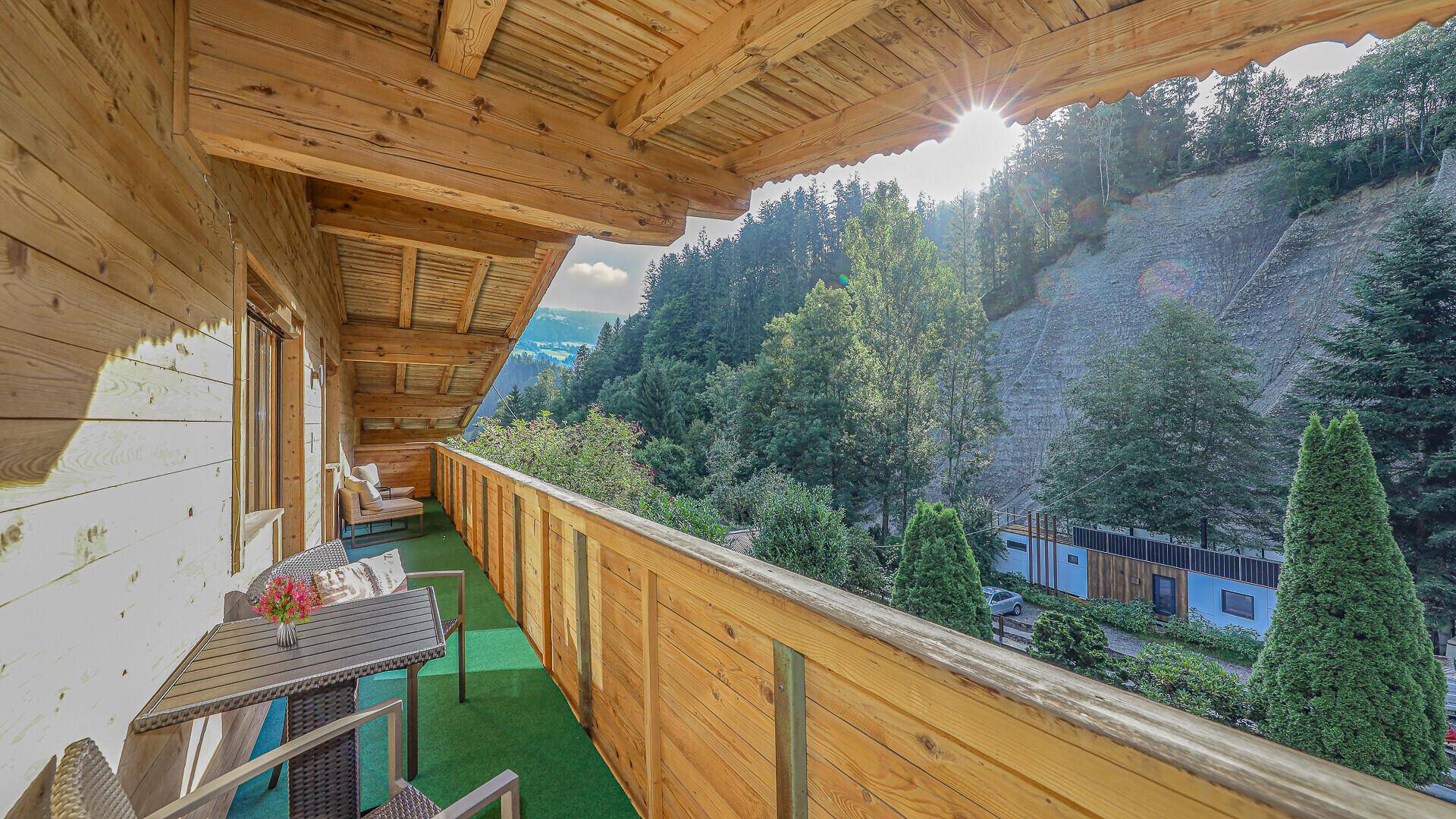 Sonniger Holzbalkon mit Sitzgelegenheiten und weitem Blick über die bewaldeten Hänge und das Tal.