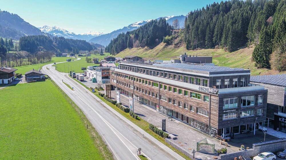 Weitläufige Luftaufnahme eines modernen Gebäudes mit Holzfassade, umgeben von grünen Feldern und Bergen.