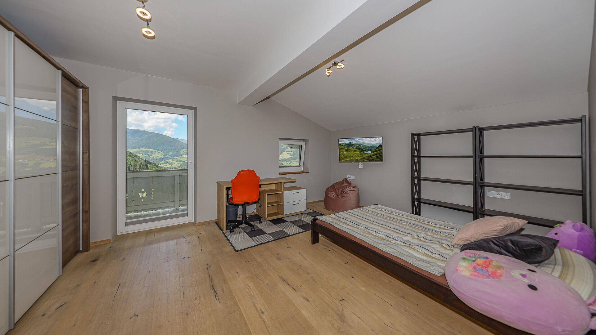 Helles Schlafzimmer mit Holzboden, Schreibtisch und Schrank, mit Balkonzugang und Bergblick.