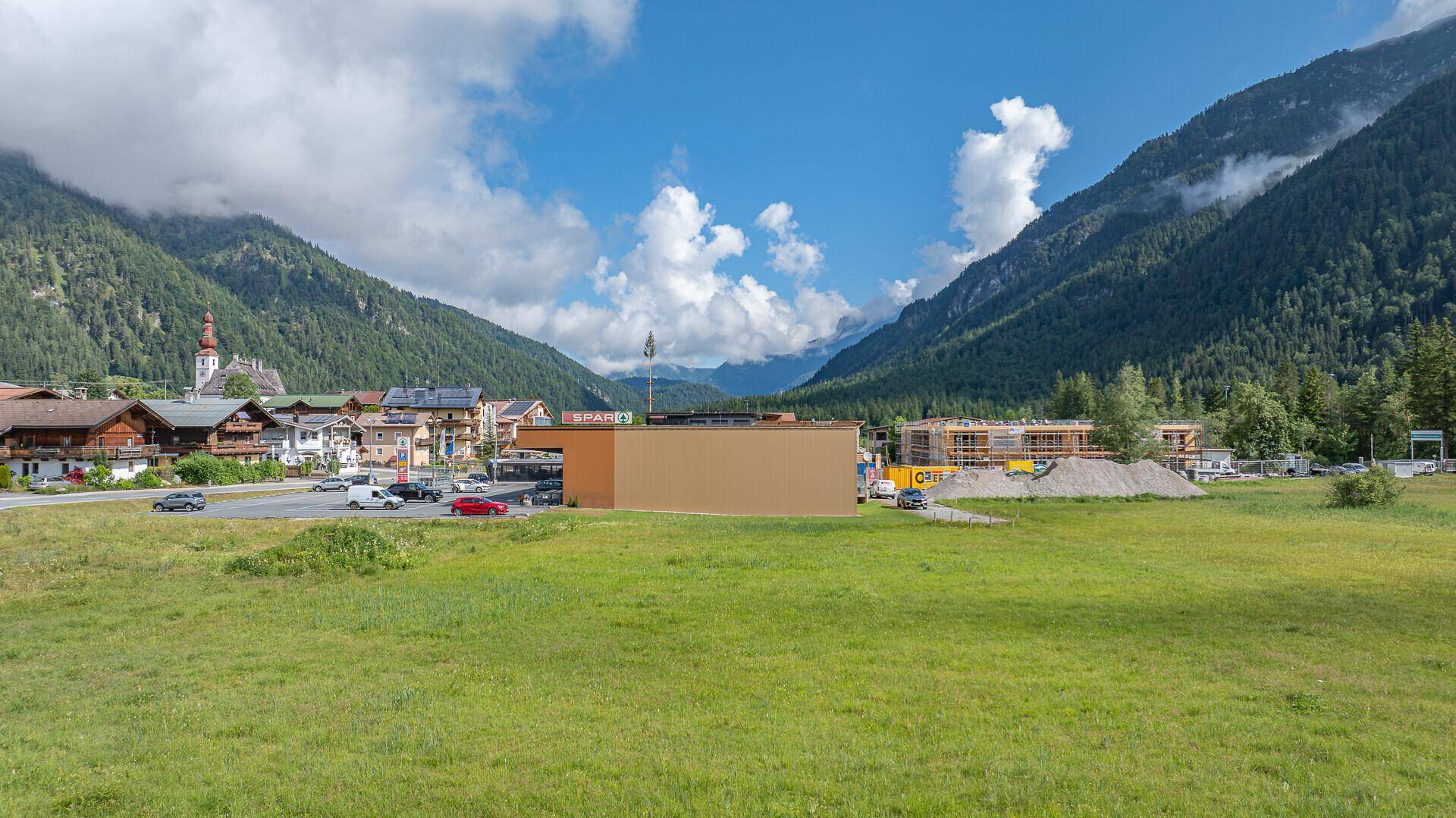 Blick auf ein Dorf mit Supermarkt und umliegenden Bergen unter einem strahlend blauen Himmel.
