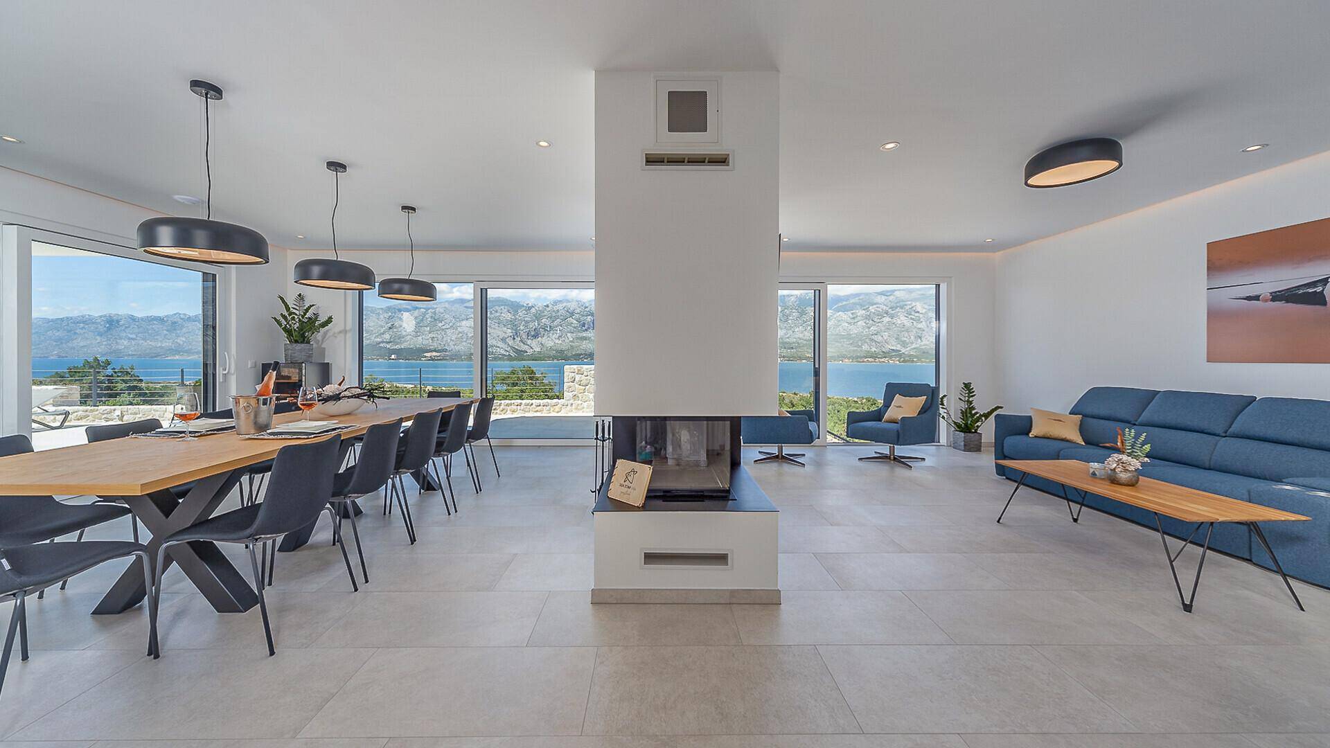 Heller Wohn- und Essbereich mit modernem Kamin, großen Fenstern und Panoramablick auf das Meer und die Berge.