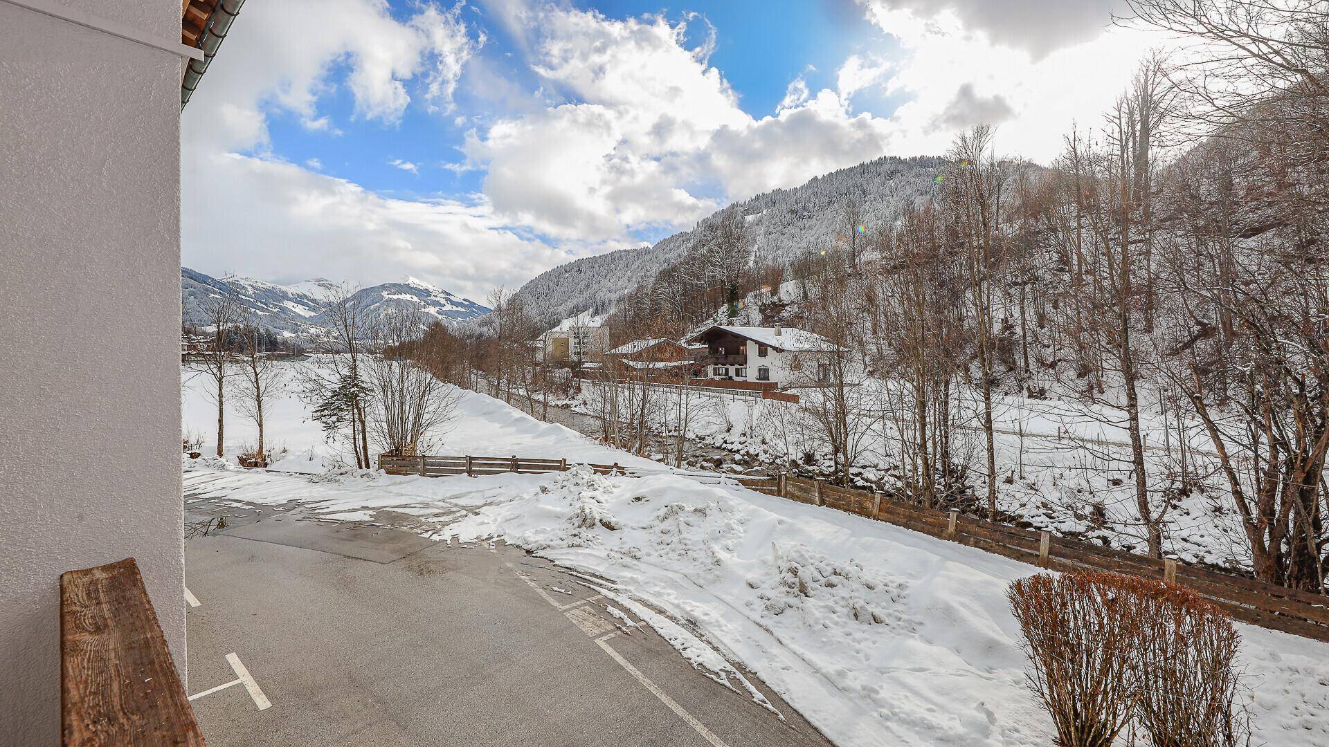 Weitläufiger Blick auf eine verschneite Berglandschaft mit Häusern, Bäumen und einer Straße im Vordergrund.