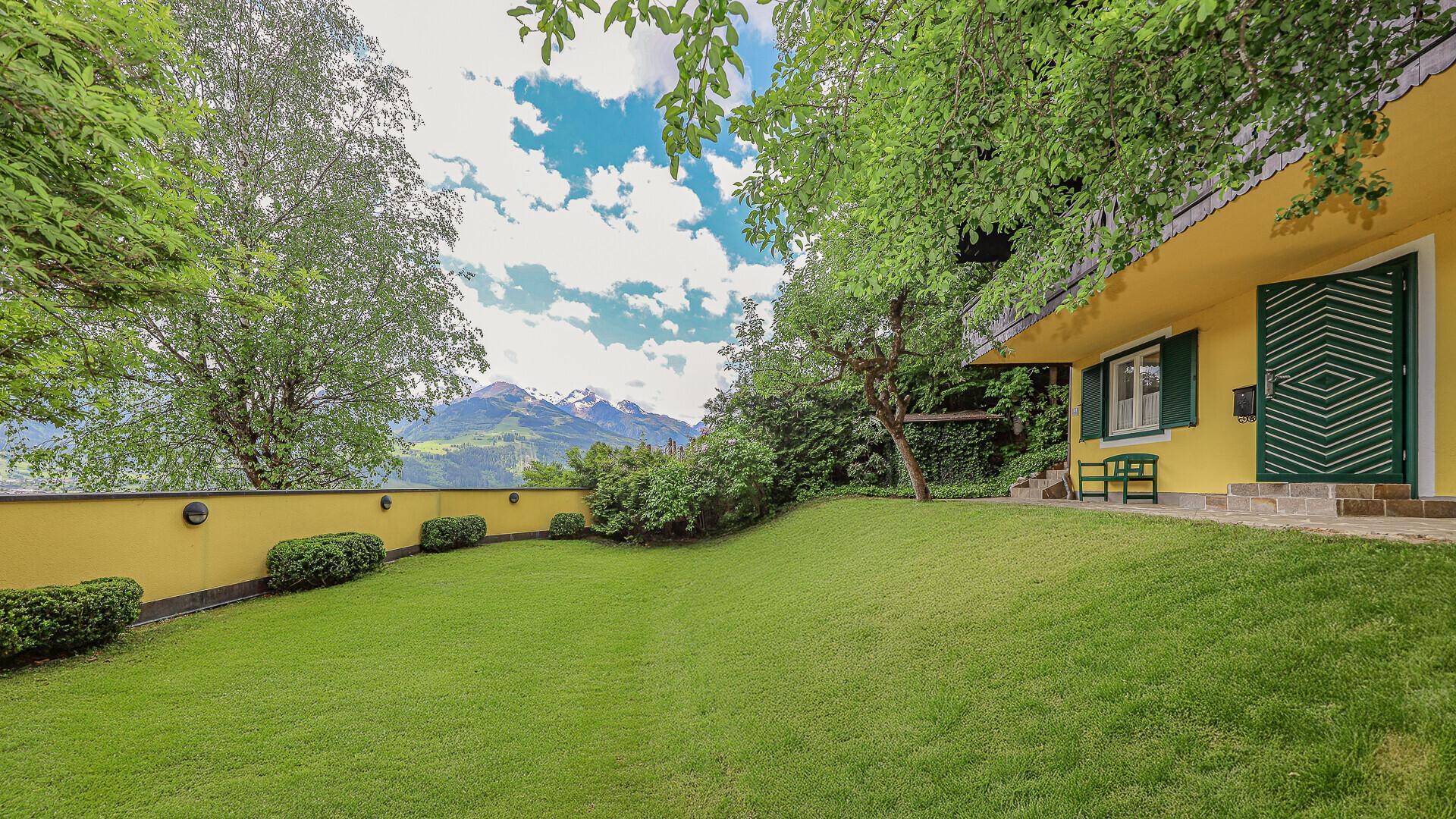 Vorderer Gartenbereich mit Rasenfläche vor dem gelben Haus und beeindruckendem Bergblick.
