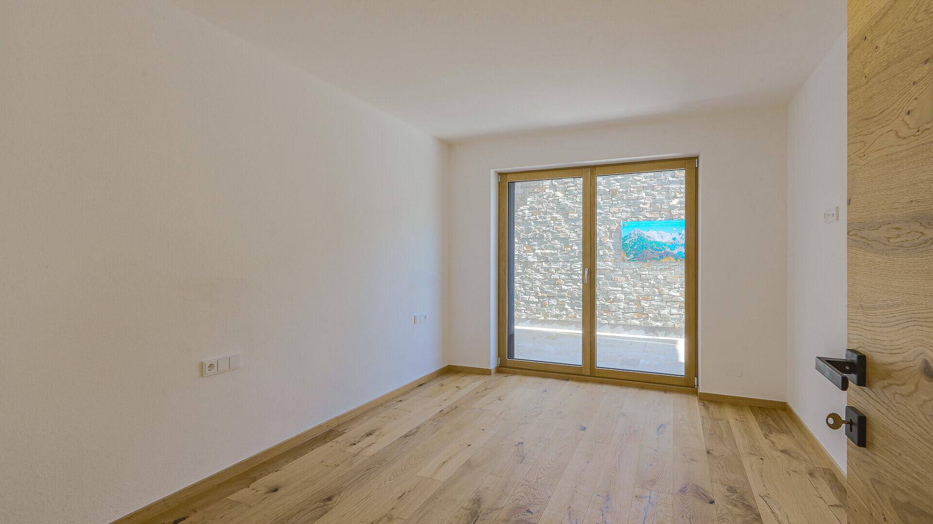 Leerer Raum mit Holzboden und großen Schiebetüren, die den Zugang zu einem Außenbereich ermöglichen.