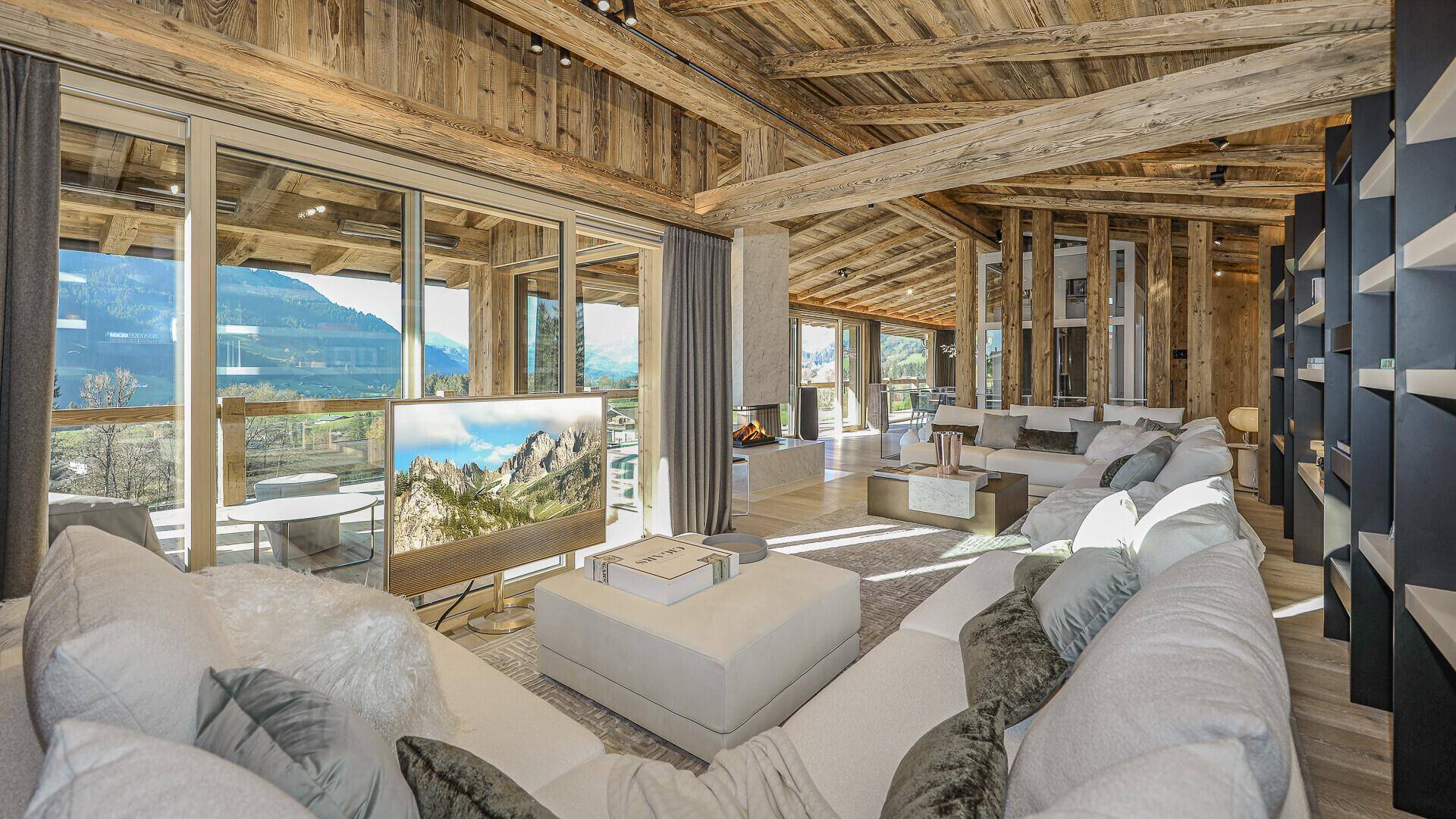 Helles Wohnzimmer mit Holzbalkendecke, großem Fernseher und Panoramablick auf die Berge.