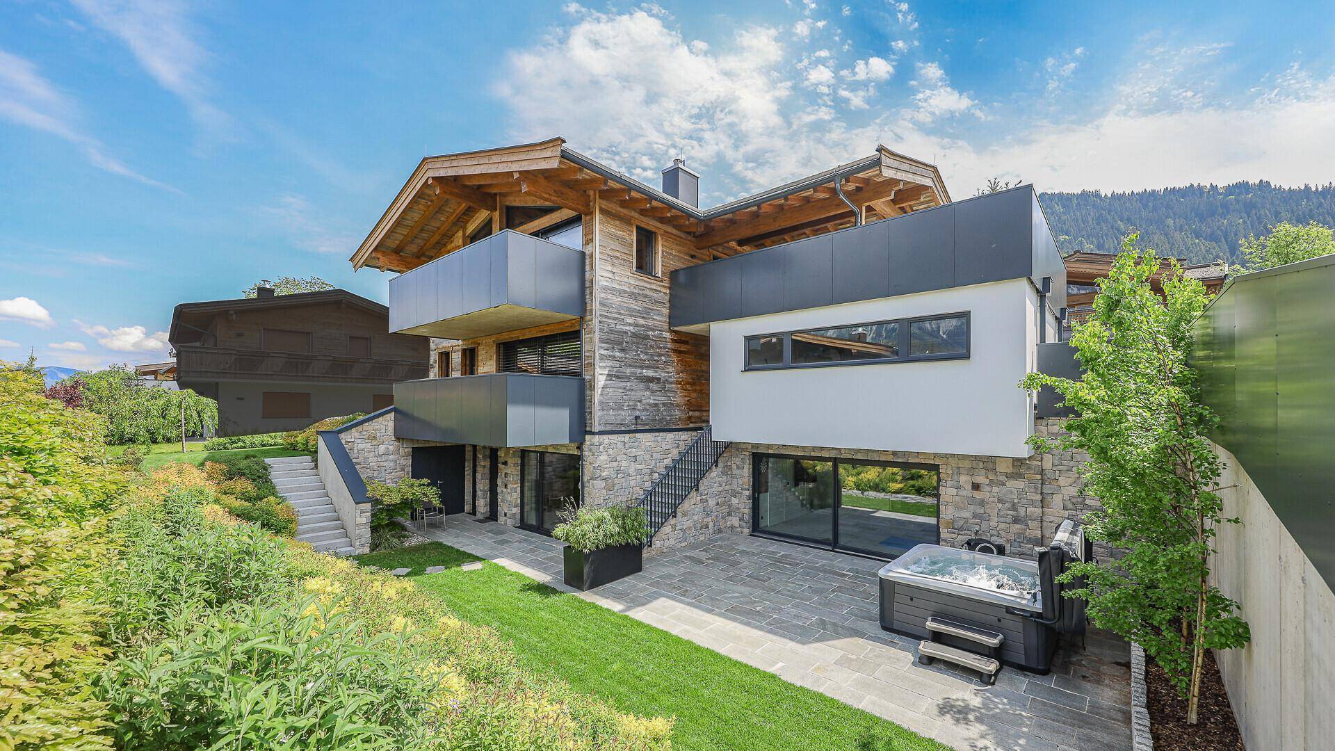 Modernes Chalet mit Holz- und Steinfassade, Garten und Whirlpool vor einer grünen Bergkulisse.