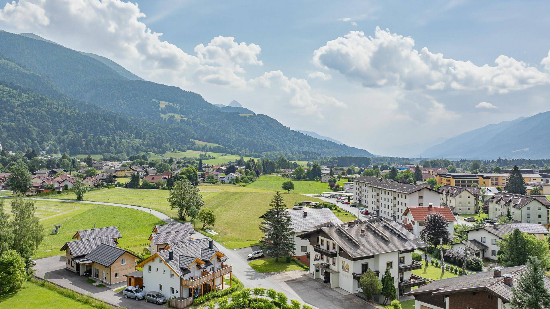 Weitläufige Luftaufnahme eines Dorfes eingebettet in eine grüne Hügellandschaft mit majestätischen Bergen im Hintergrund.