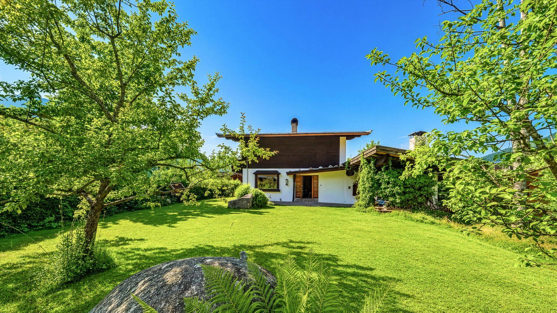 Großer, sonniger Garten hinter dem Chalet mit Rasenfläche, Bäumen und Felsen.