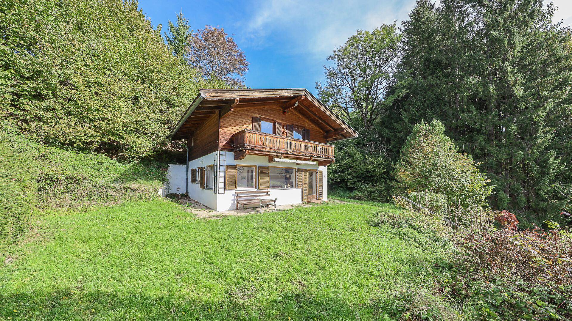 Alpines Holzhaus mit Balkon und großem Rasenbereich, umgeben von hohen Bäumen.
