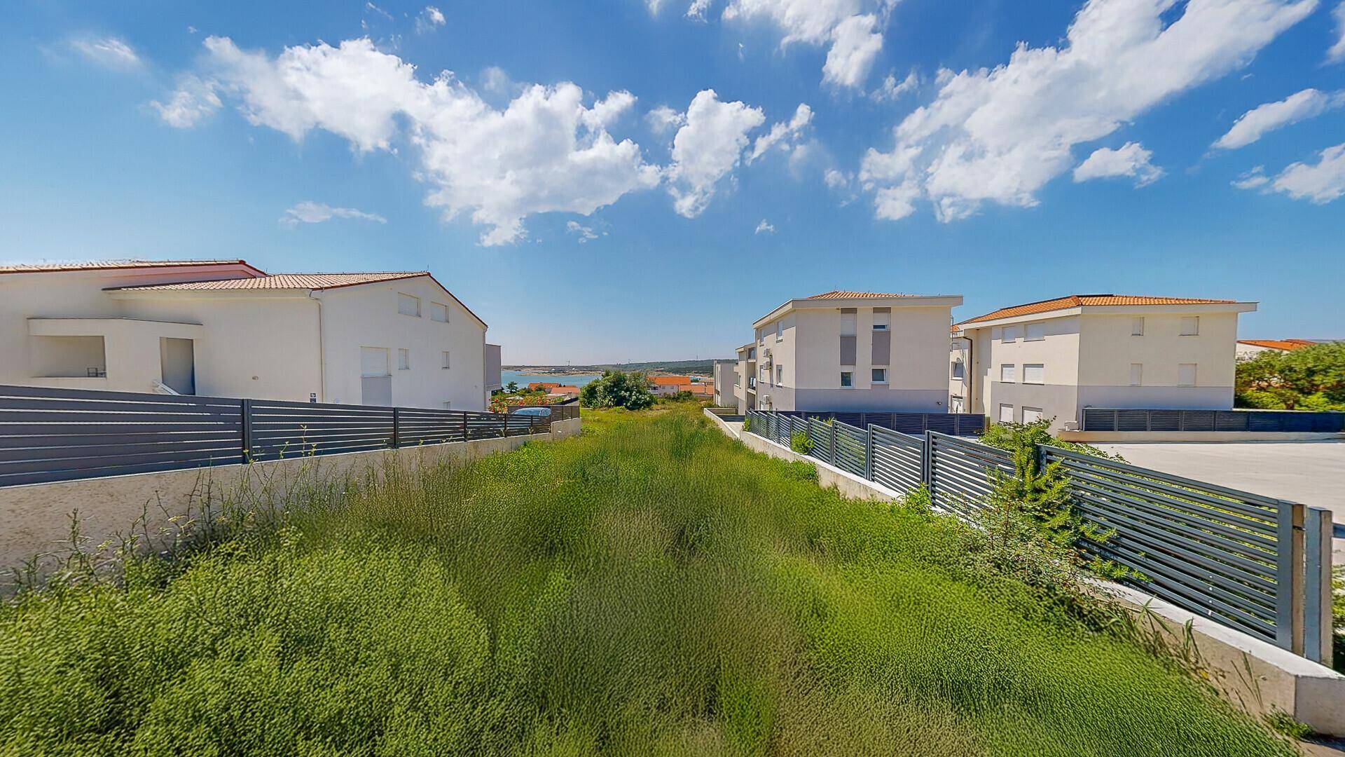 Grüne Fläche eines Grundstücks zwischen modernen Gebäuden, mit Blick auf das Meer am Horizont.