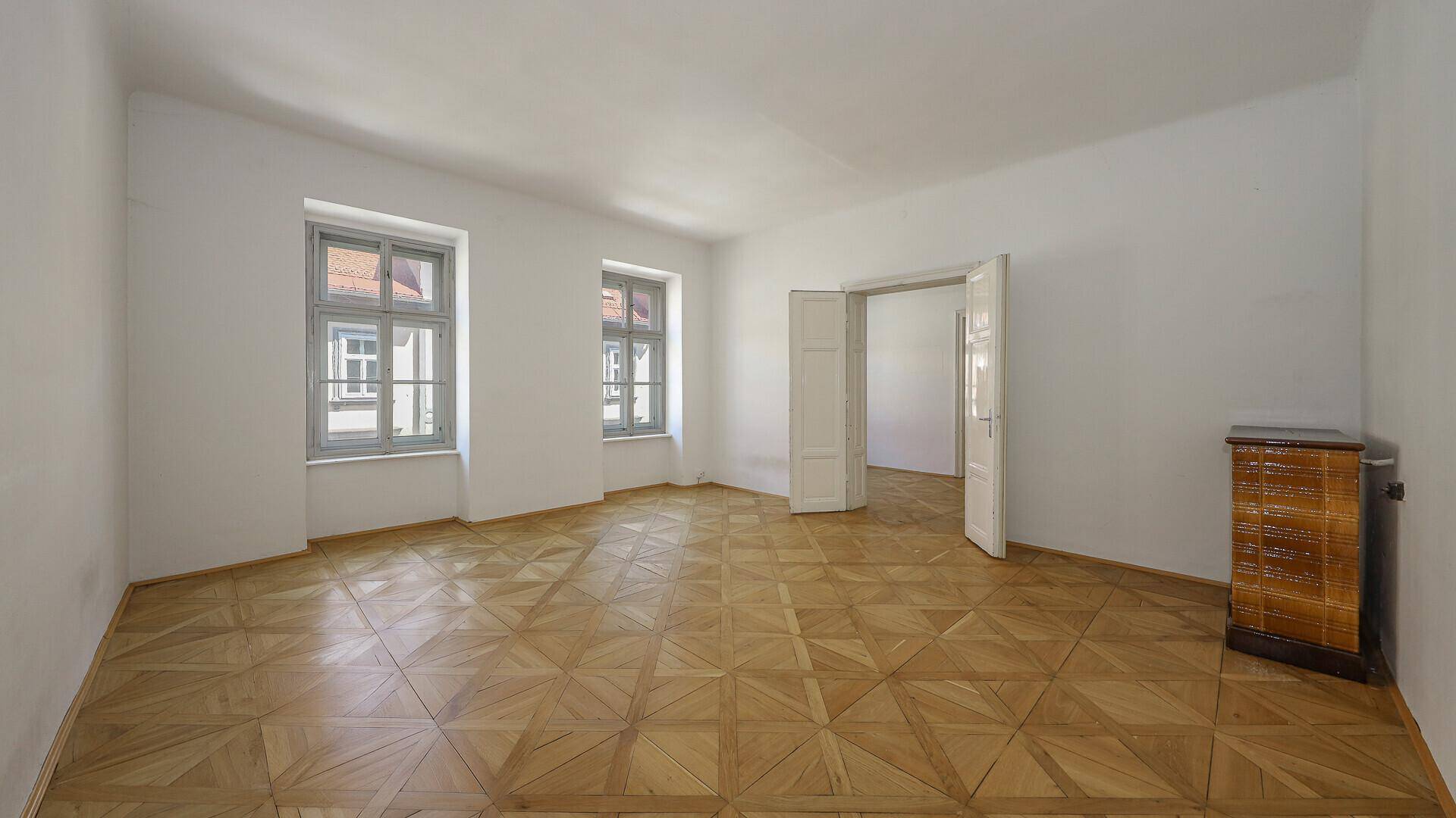 Helles Zimmer mit Fischgrätparkett, hohen Decken und Zugang zu weiteren Räumen.