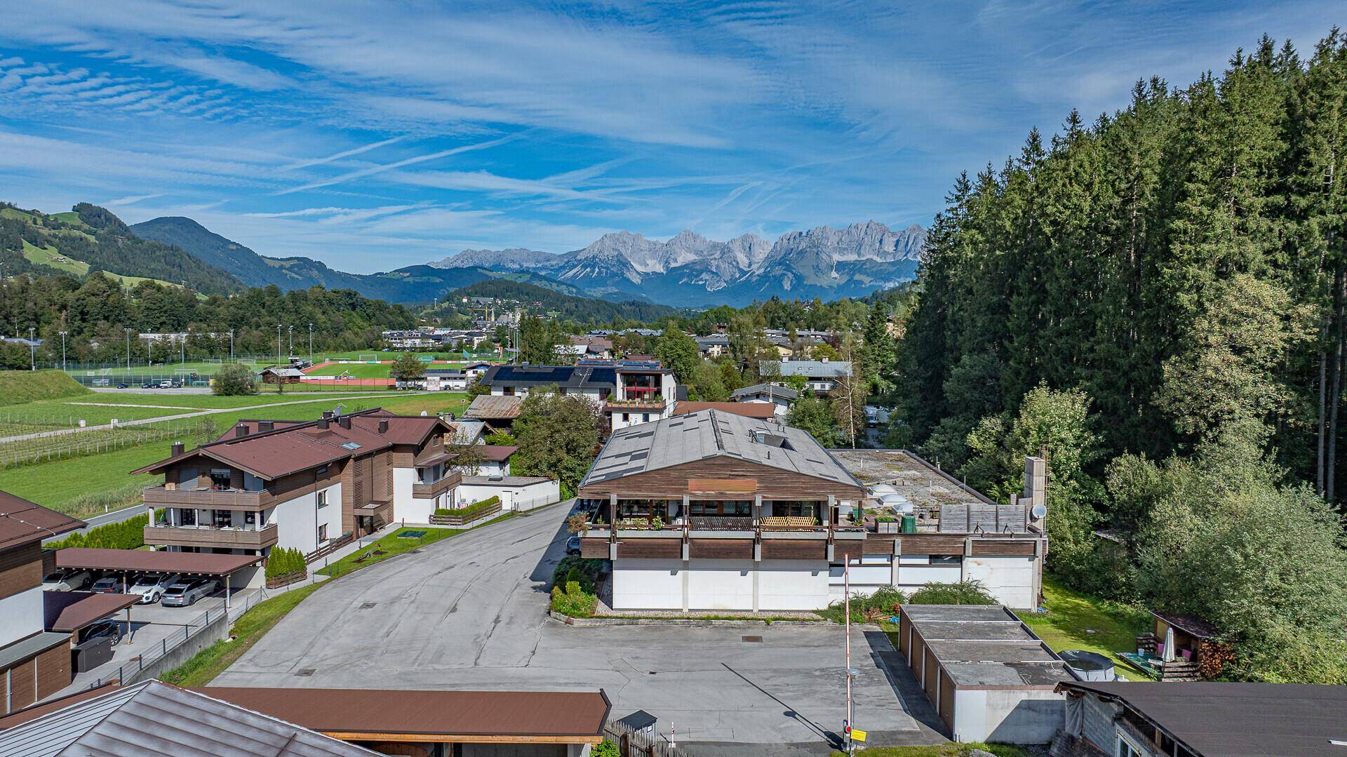 Panoramablick auf das Gebäude mit Flachdach, umgeben von Sportanlagen und alpiner Kulisse.
