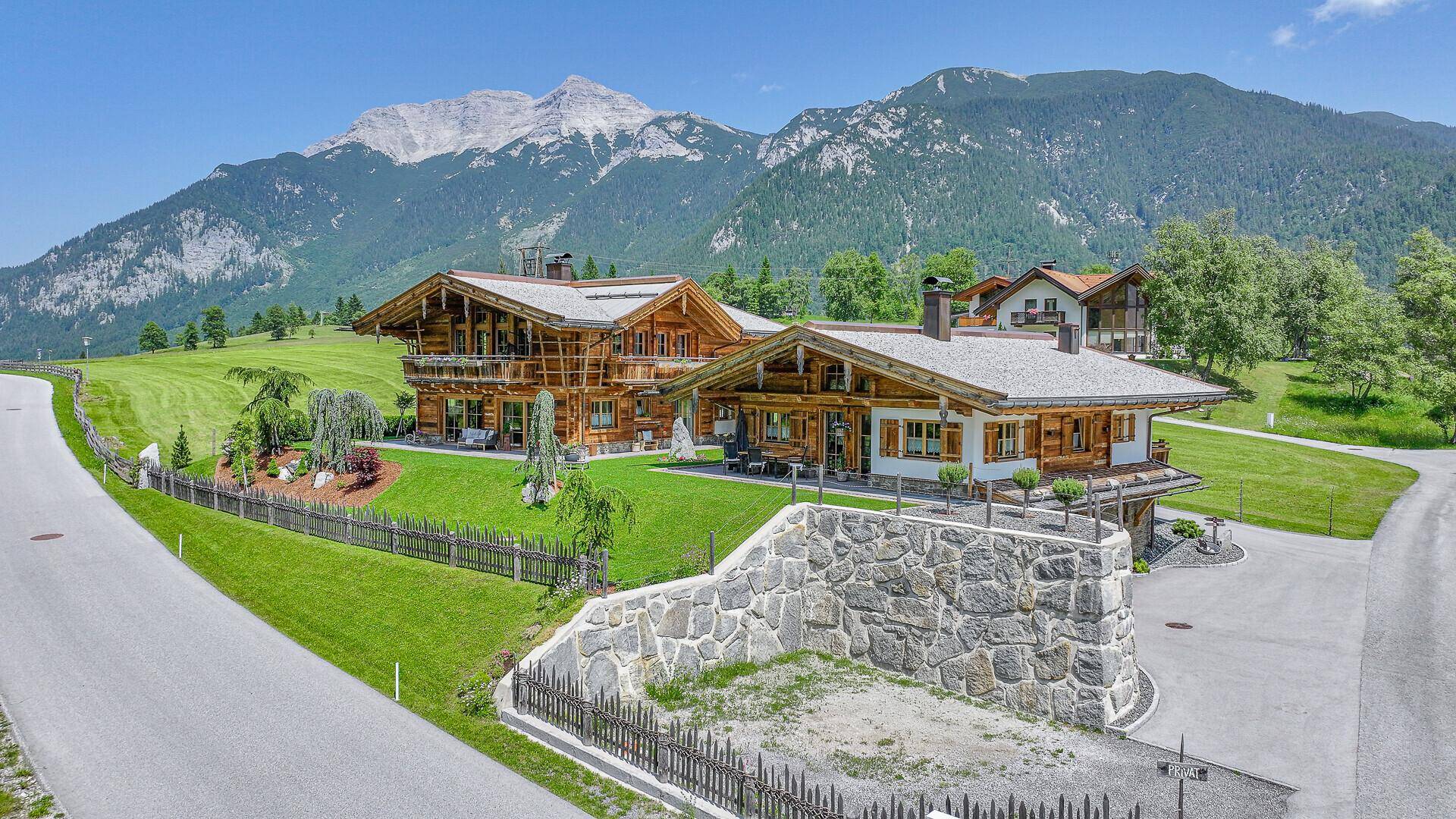 Zwei moderne Chalets mit Holzbalkonen und gepflegten Gärten vor einer beeindruckenden Bergkulisse.
