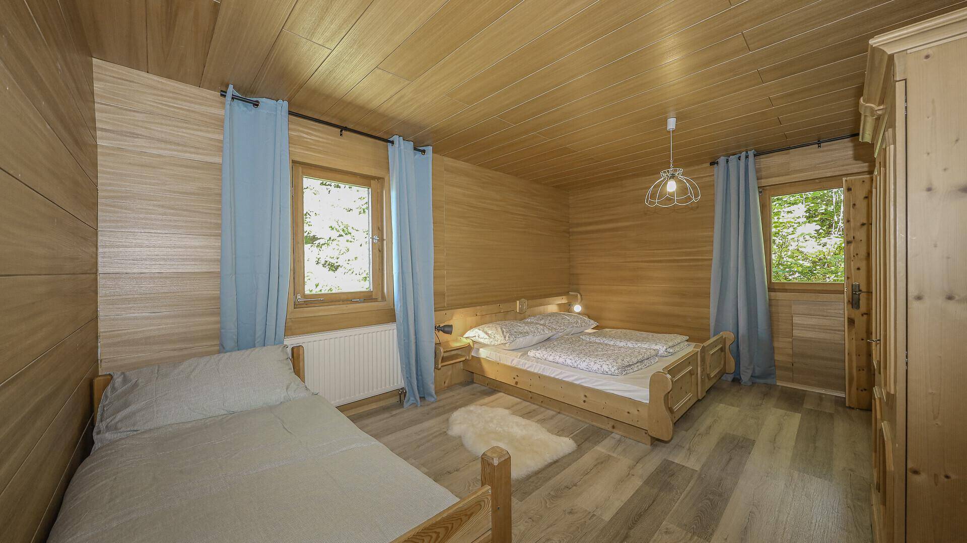 Gemütliches Schlafzimmer mit Holzwänden, Holzboden und zwei Fenstern mit Blick ins Grüne.