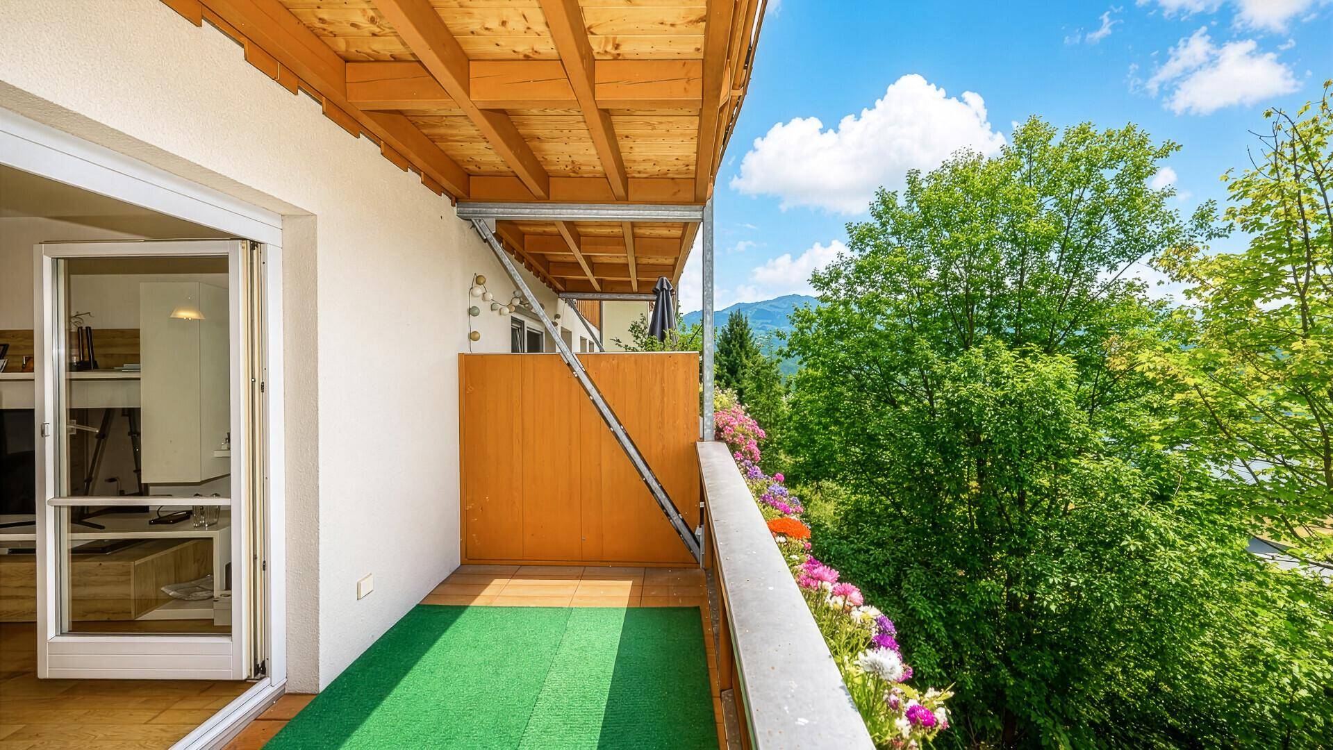 Sonniger Balkon mit Kunstrasen, Holzdach und Blick auf grüne Bäume und Berge.