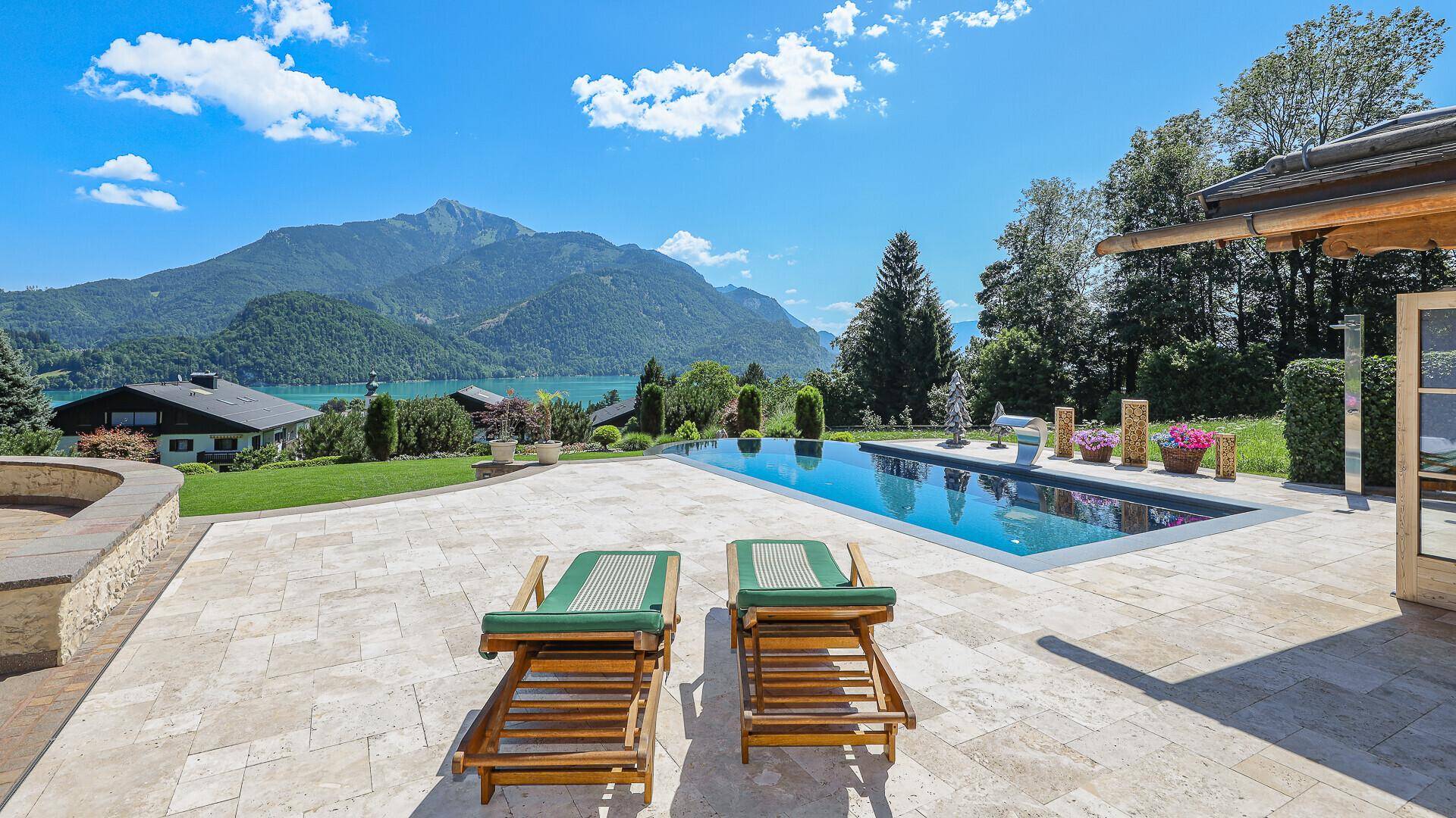 Sonniger Poolbereich mit Liegestühlen und weitem Blick auf den See und die Berge.
