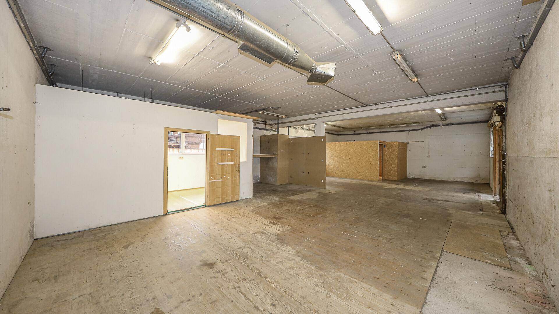 Geräumiger Lagerraum mit Betonboden, sichtbaren Lüftungsrohren und provisorischen Trennwänden aus Holz.