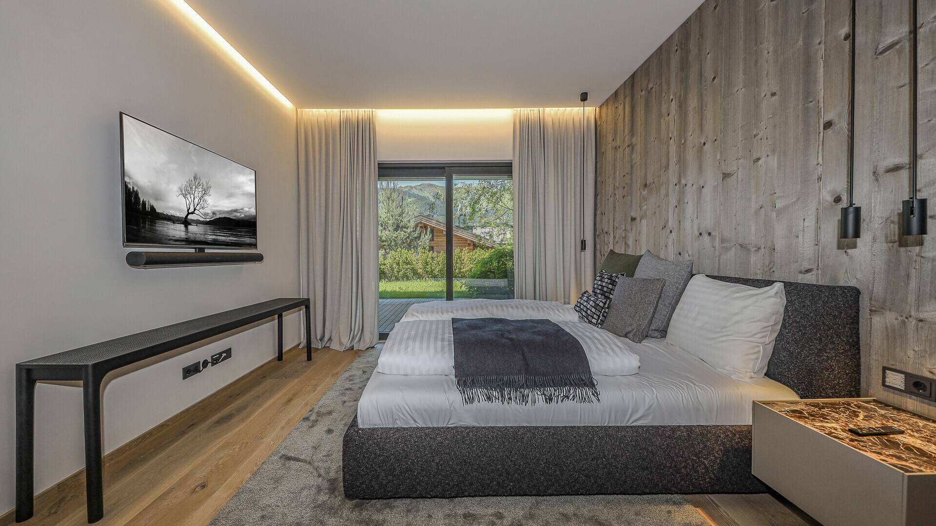 Modernes Schlafzimmer mit Holzwand, großem Bett und Zugang zu einer Terrasse mit Gartenblick.