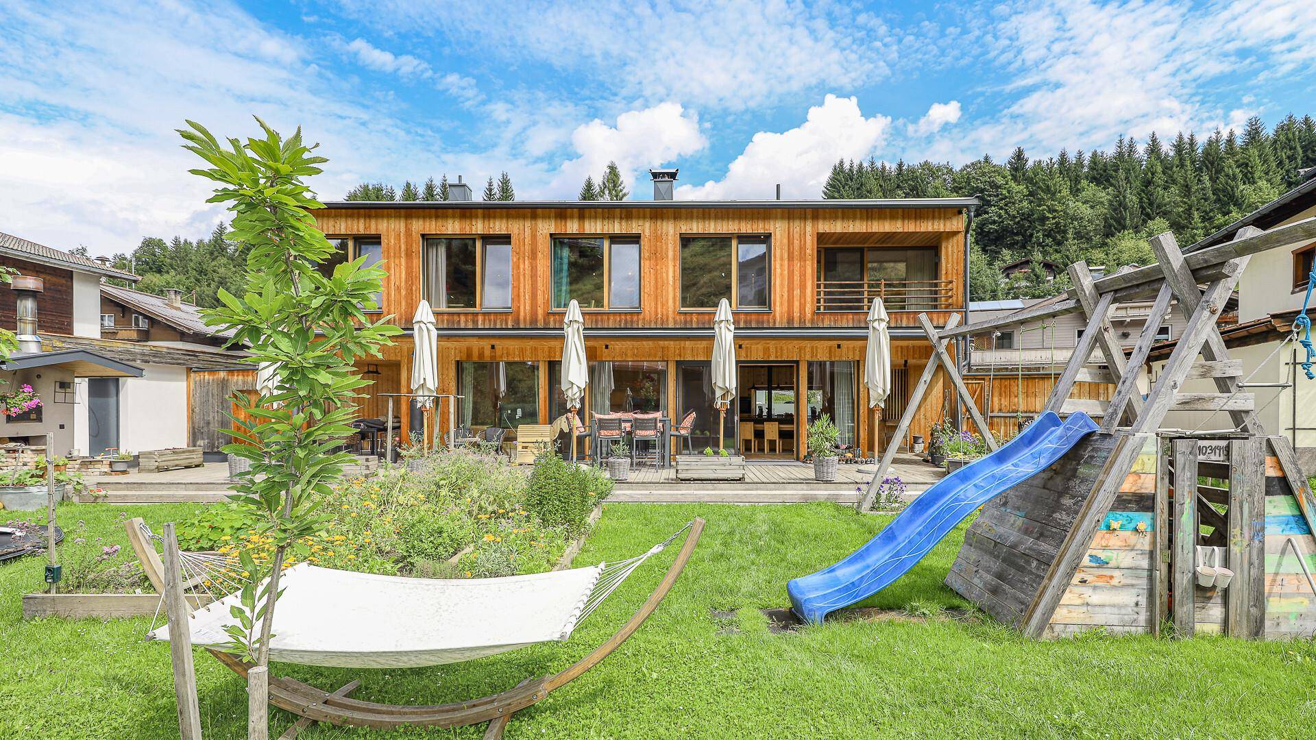 Großer Garten mit Spielplatz, Hängematte und Blick auf die moderne Holzfassade des Hauses.