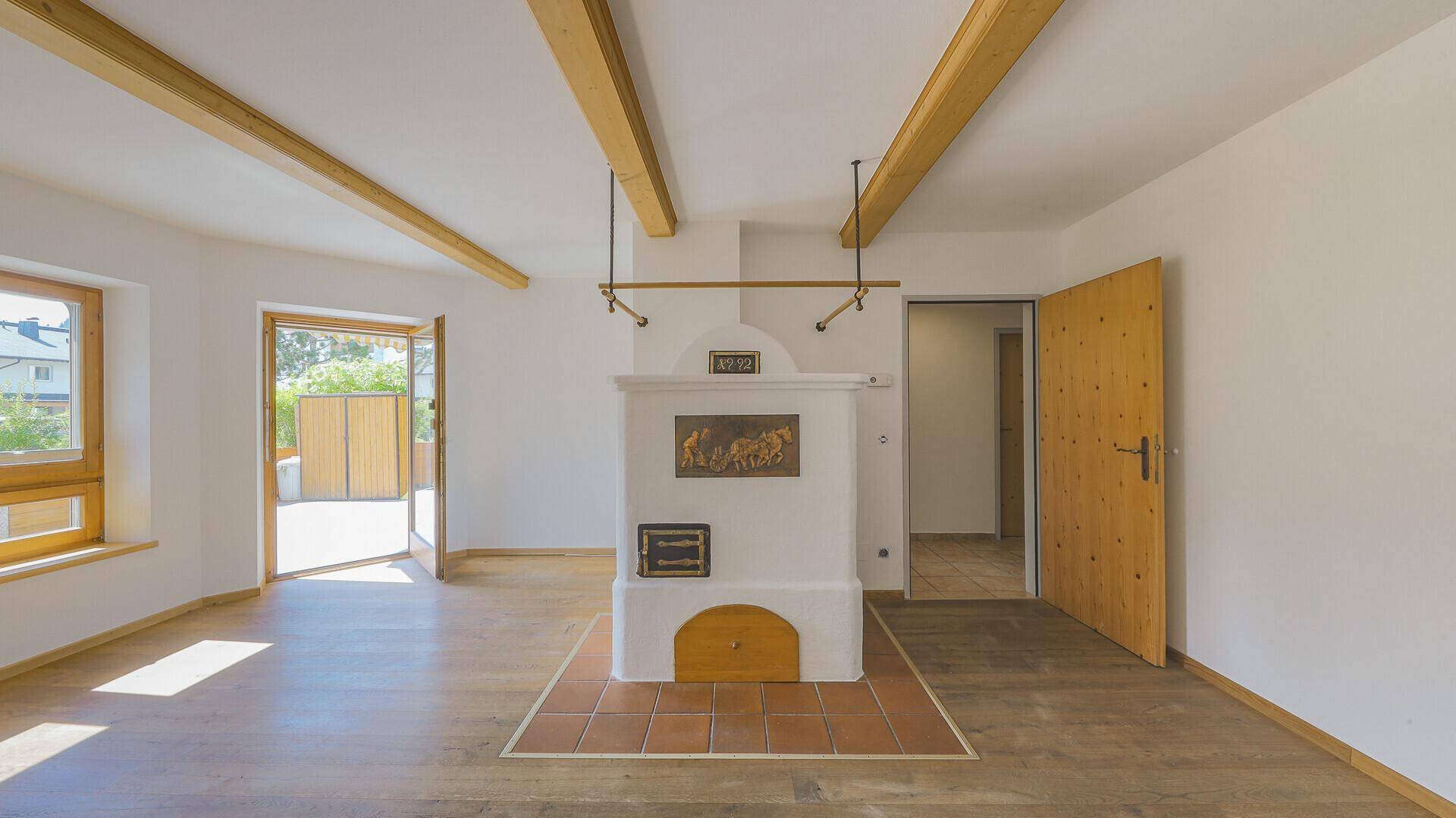 Helles Wohnzimmer mit Kachelofen, Holzbalken und Holzboden, mit Zugang zu einem weiteren Raum.