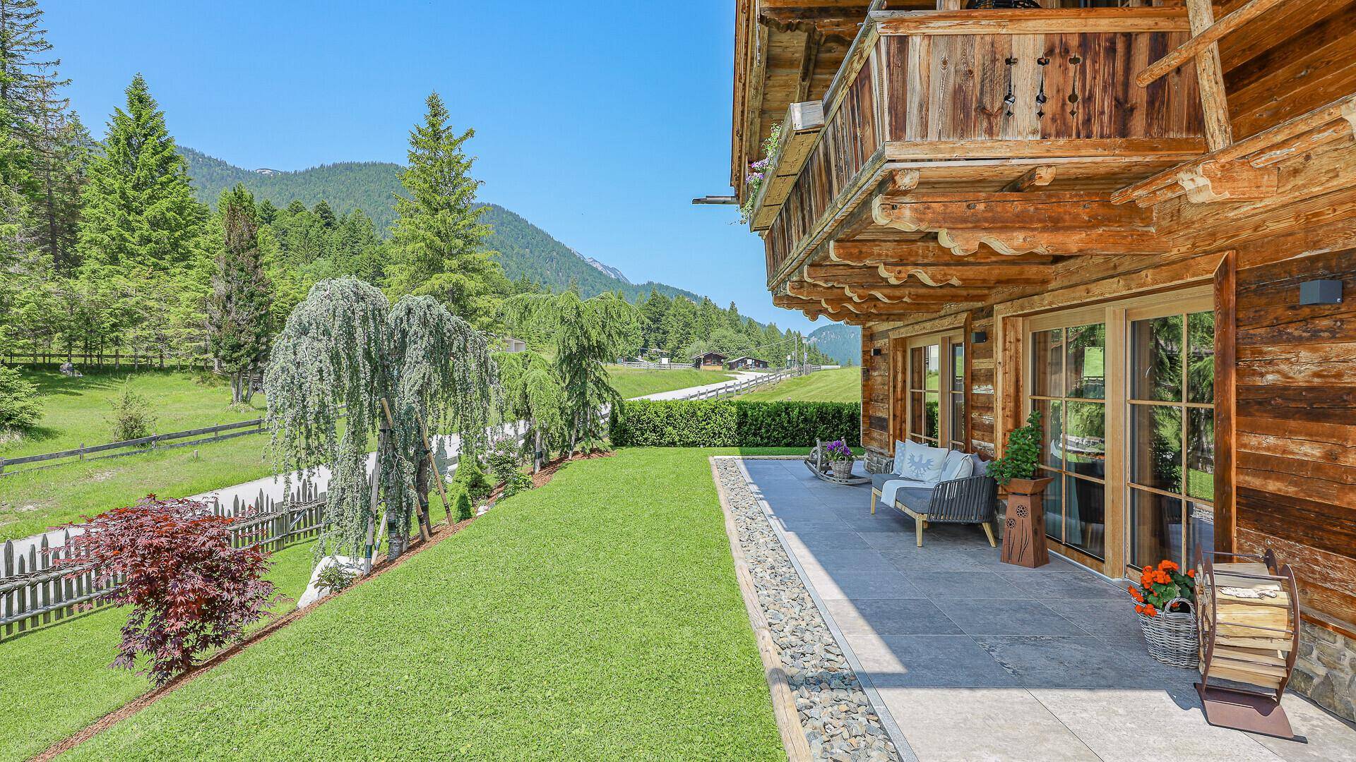 Sonnige Terrasse mit bequemen Gartenmöbeln und gepflegtem Rasen, umgeben von Bäumen.
