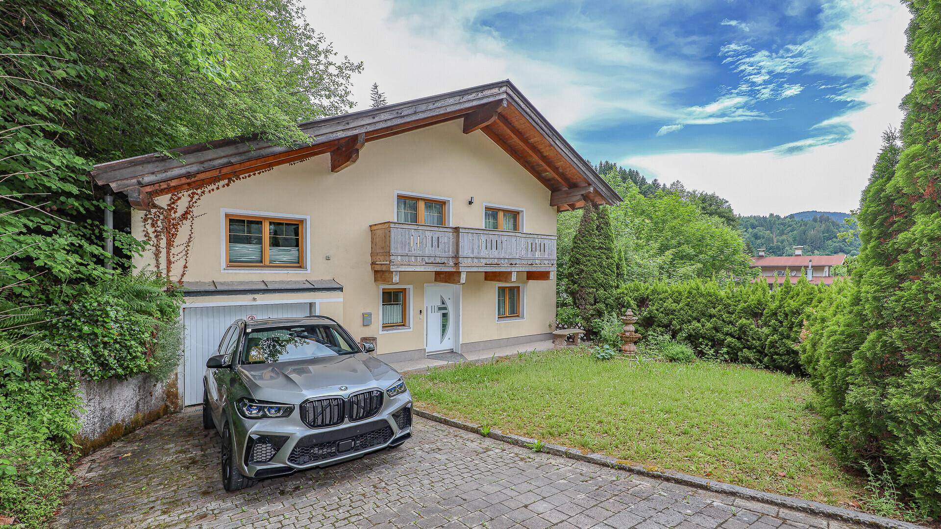 Einfamilienhaus mit Garage, gepflasterter Auffahrt und großem Vorgarten umgeben von Bäumen.