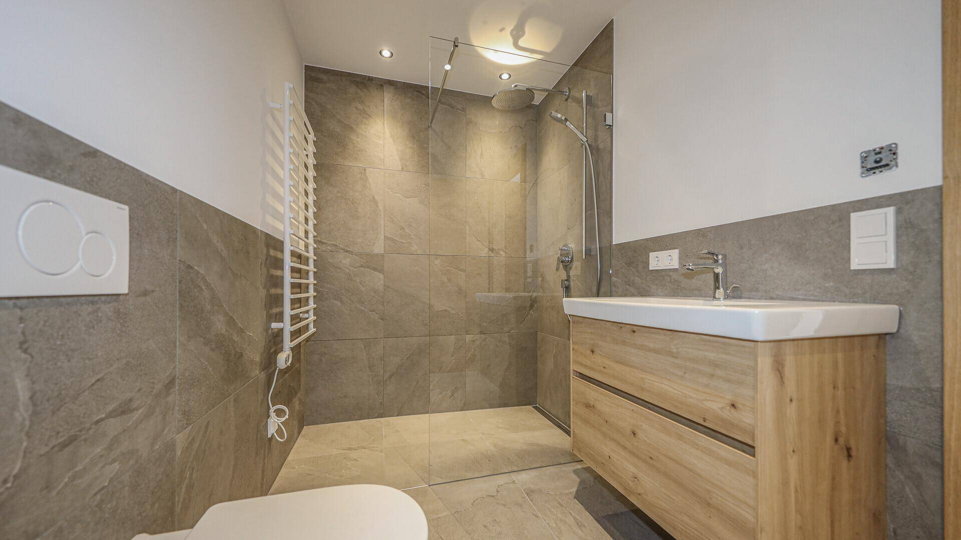 Modernes Badezimmer mit grauen Fliesen, Holzwaschtisch, wandhängendem WC und begehbarer Dusche.