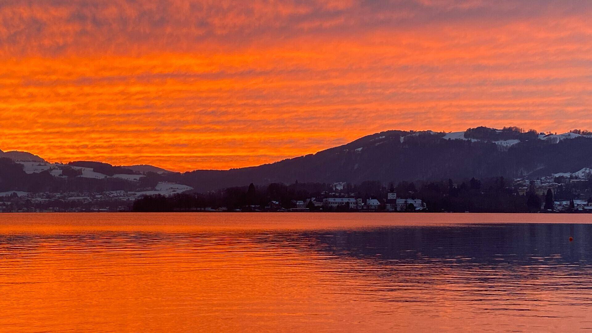 Atemberaubender Sonnenaufgang über einem See mit orangefarbenem Himmel und schneebedeckten Bergen im Hintergrund.