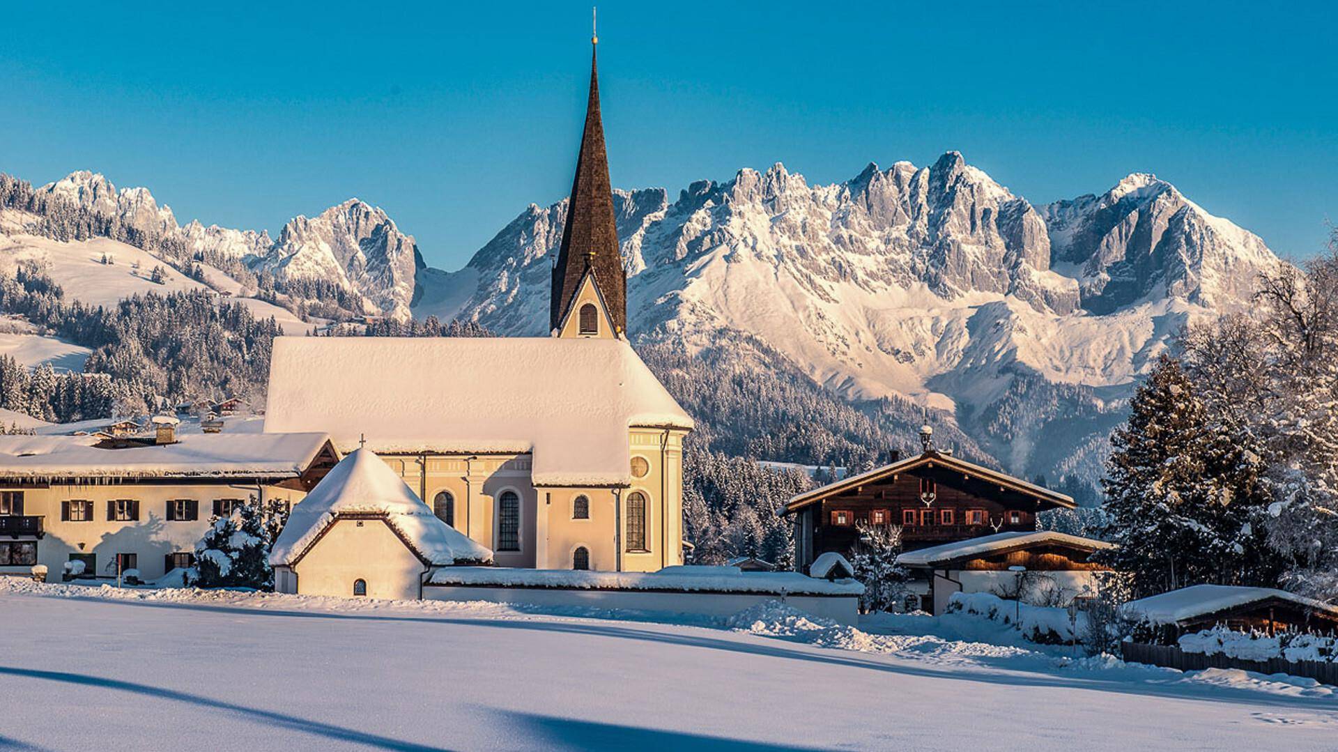 Verschneite Kirche und traditionelle Häuser vor einer majestätischen Berglandschaft im Winter.