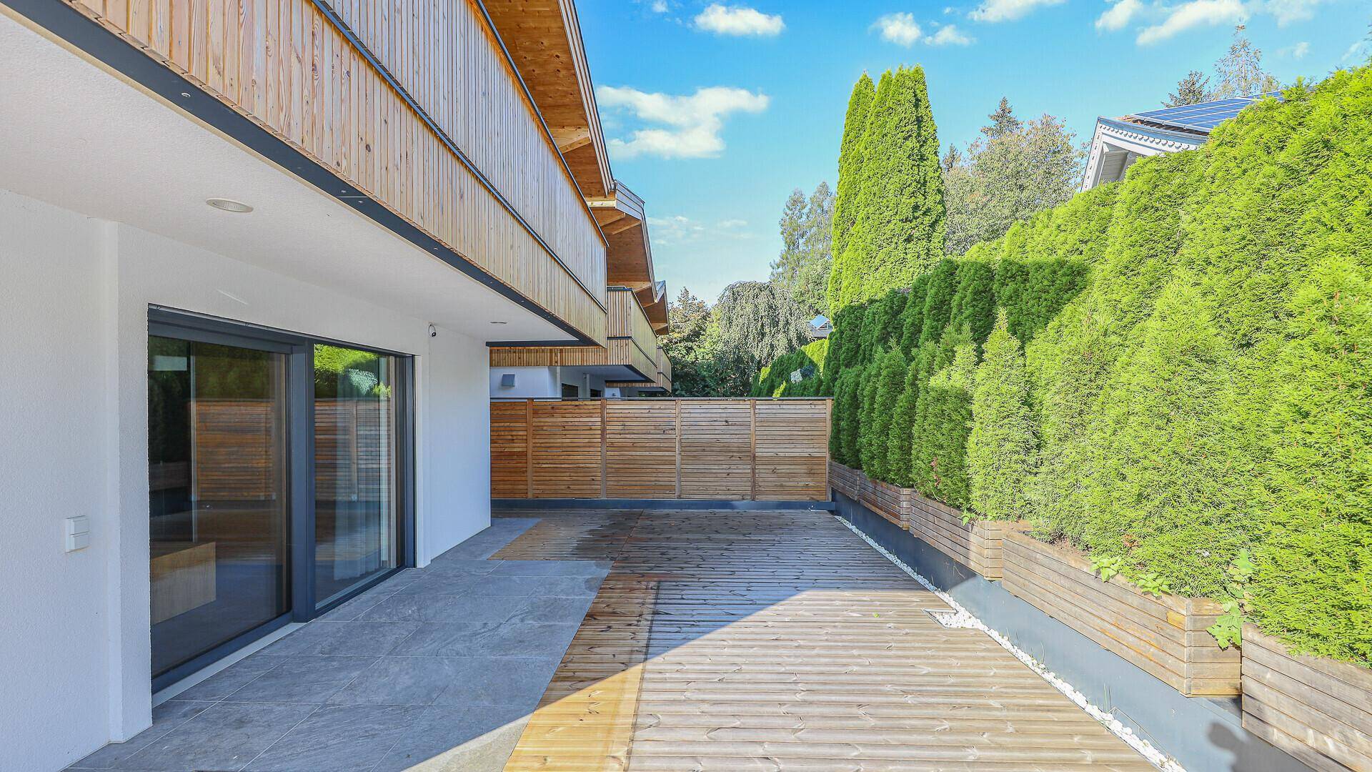 Geräumige Terrasse mit Holzboden und Blick auf eine grüne Hecke und den Garten.