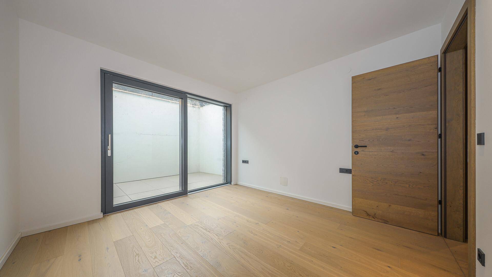 Helles Zimmer mit Holzboden und Schiebetür, die zu einem Lichtschacht oder kleinen Außenbereich führt.