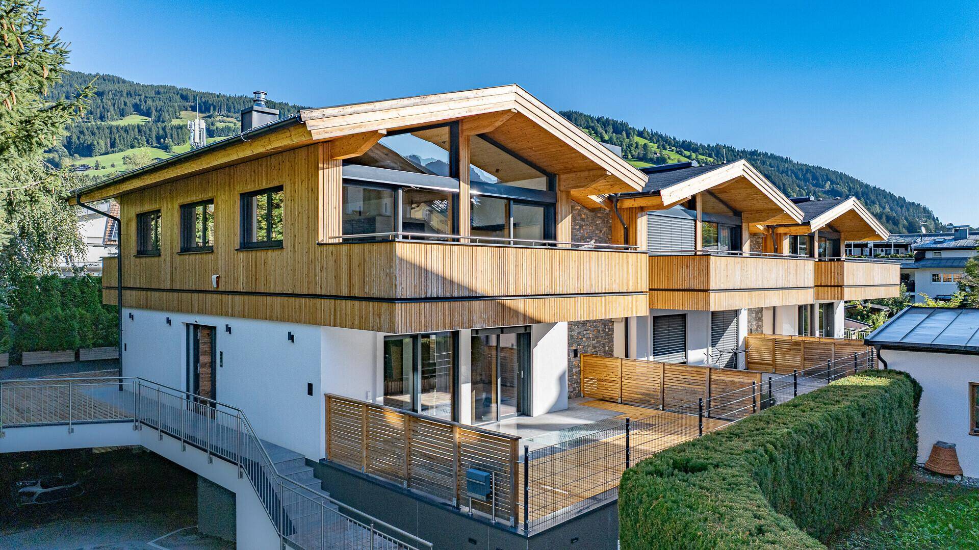 Modernes Mehrfamilienhaus mit Holzfassade, großen Fenstern und Balkonen in alpiner Umgebung.