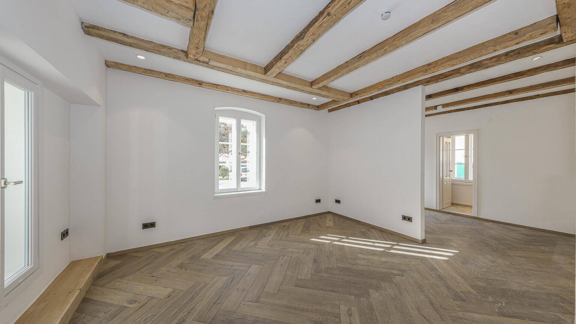 Geräumiges Zimmer mit Fischgrätparkett, weißen Wänden und Holzbalkendecke.