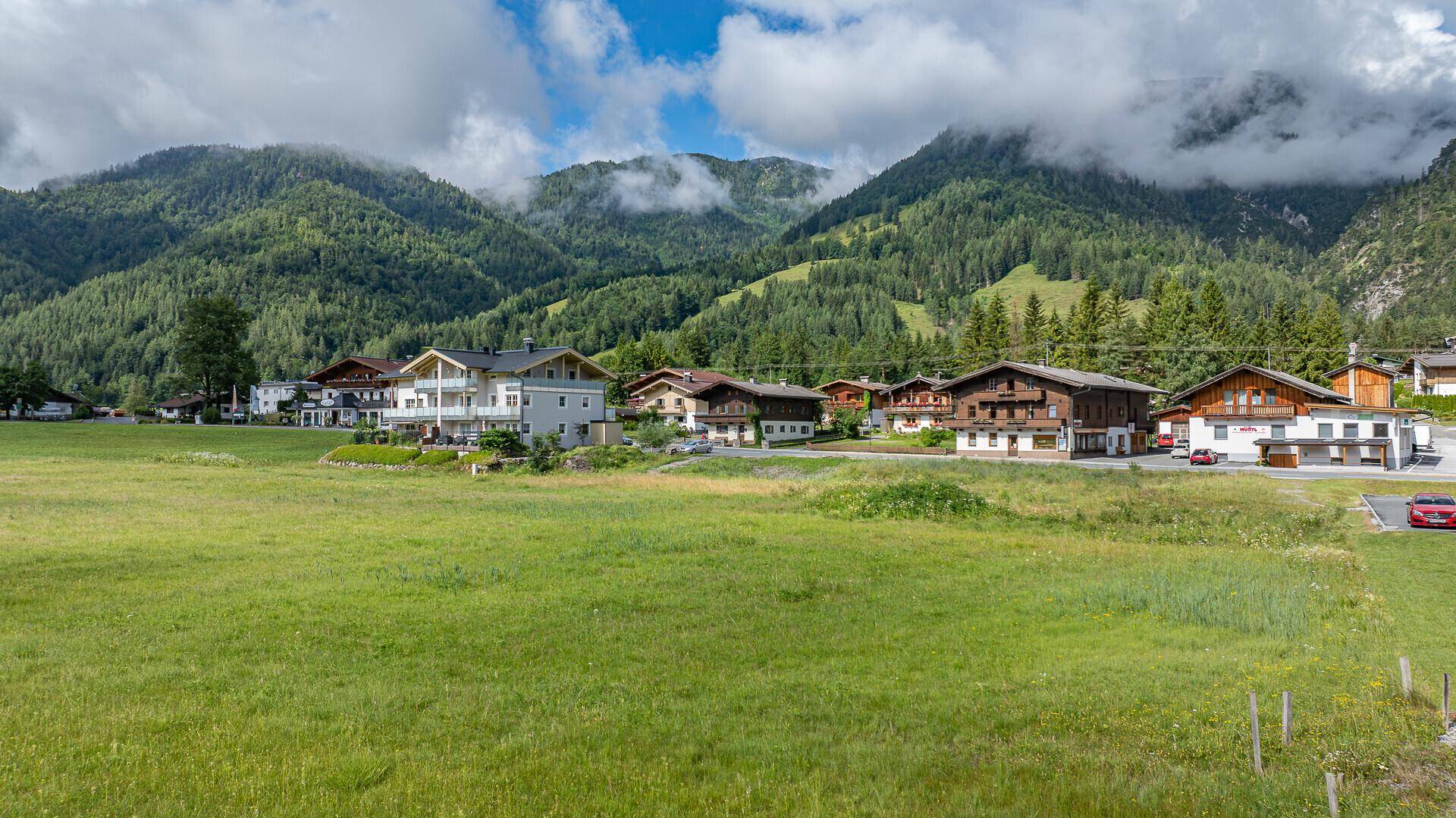 Wohngebiet in einem grünen Tal, umgeben von bewaldeten Bergen und weiten Wiesen.