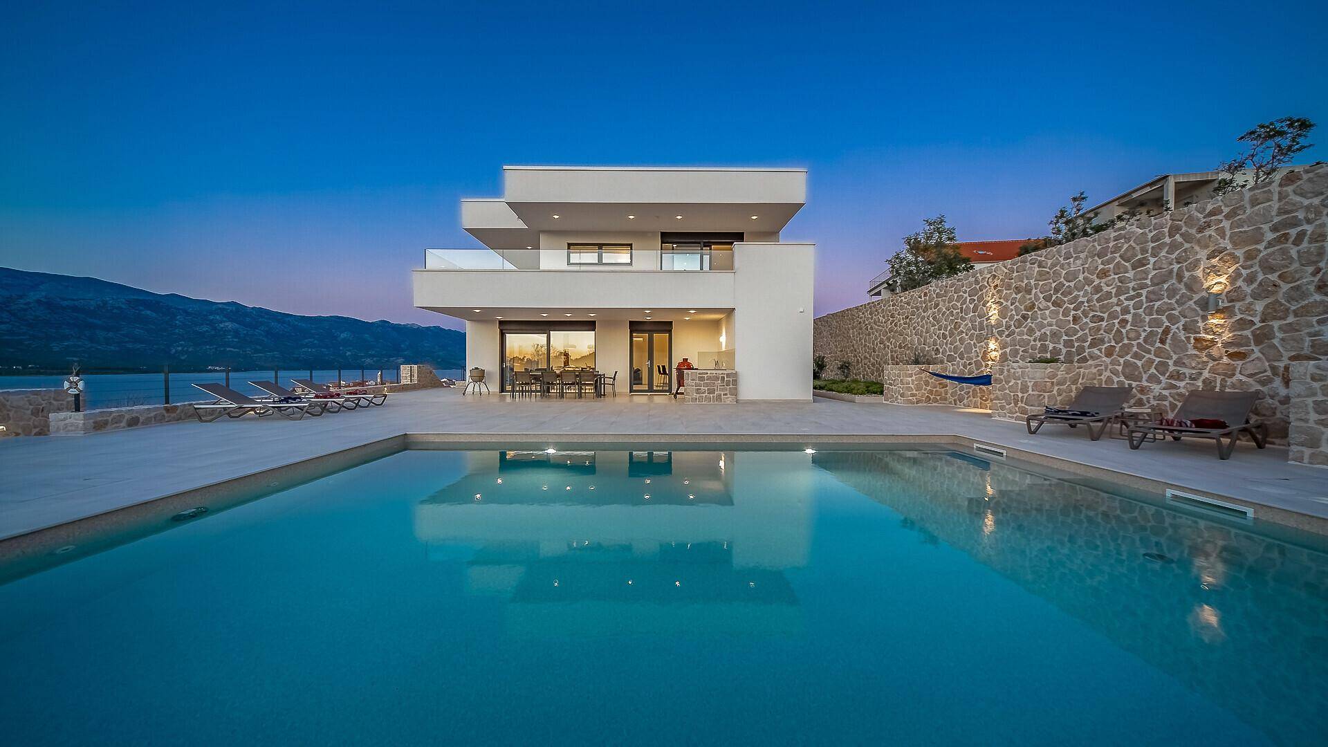 Elegante Villa bei Sonnenuntergang mit beleuchtetem Pool und Steinmauern, umgeben von Natur.
