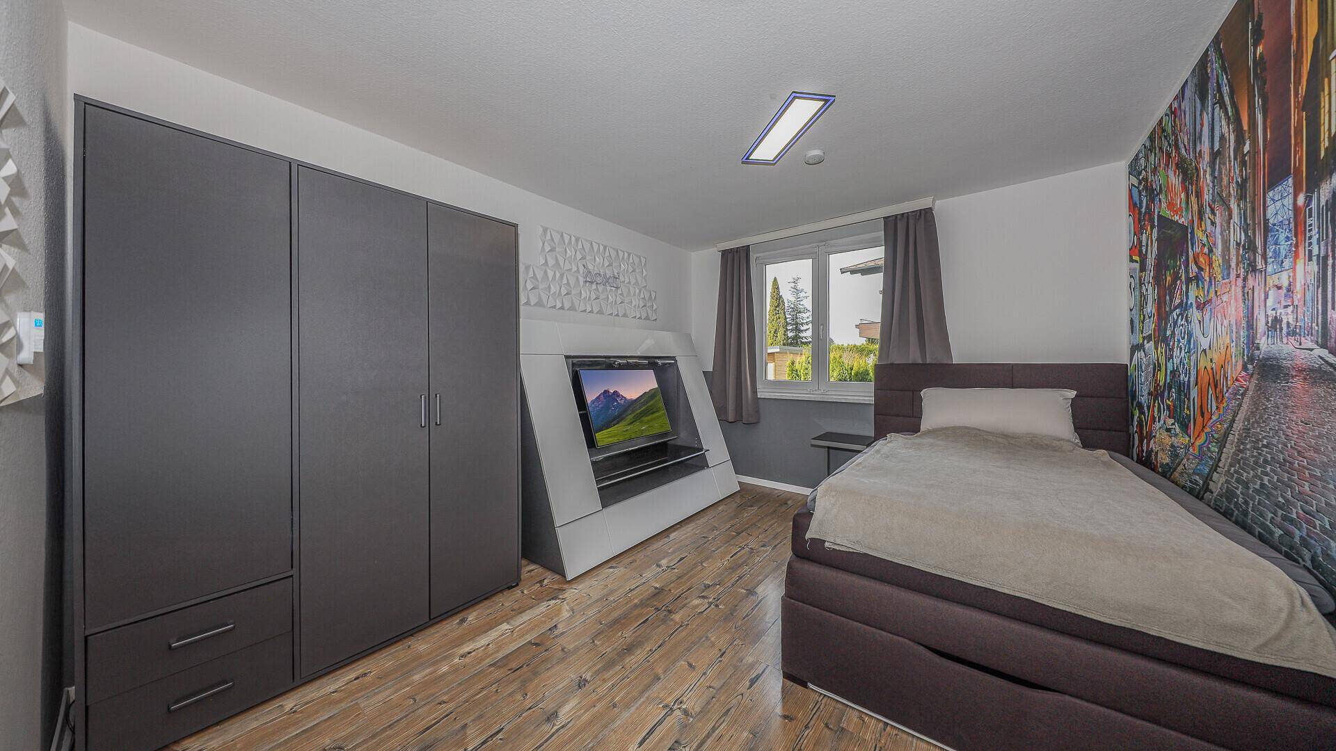 Jugendliches Schlafzimmer mit Einzelbett, dunklem Kleiderschrank und farbenfroher Wandgestaltung.
