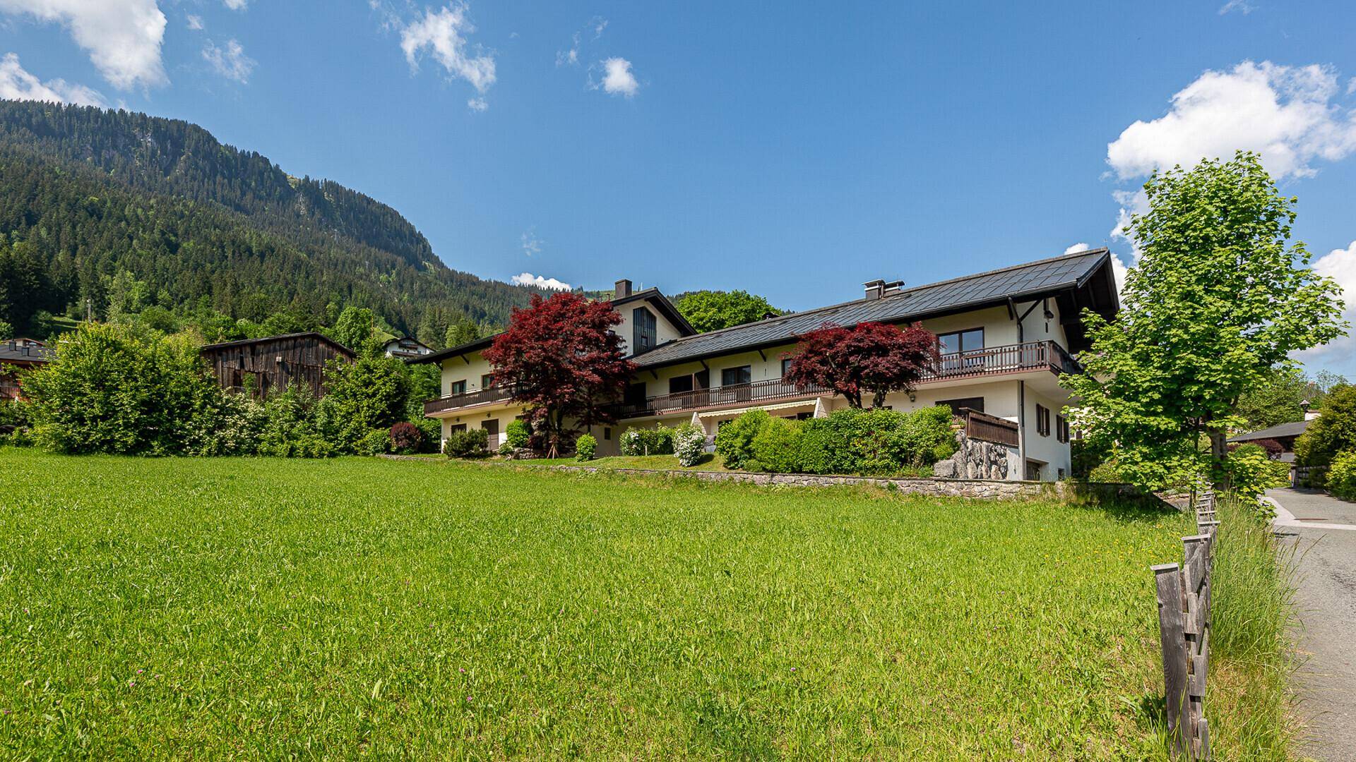 Großes Wohnhaus mit weitläufiger Rasenfläche und beeindruckendem Bergpanorama unter blauem Himmel.