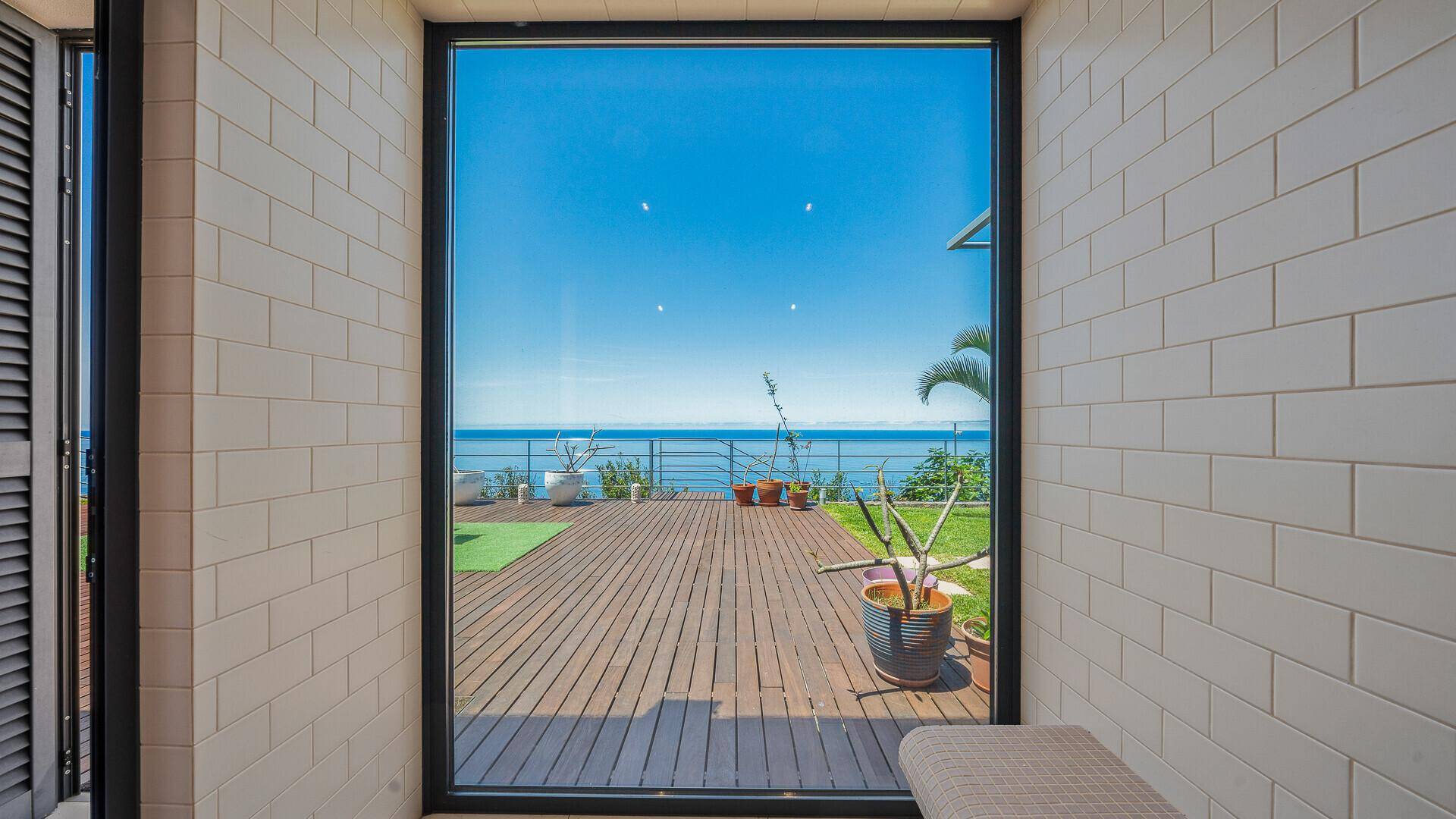 Innenraum mit großen Fenstern, die einen weiten Blick auf eine Holzterrasse und das Meer bieten.