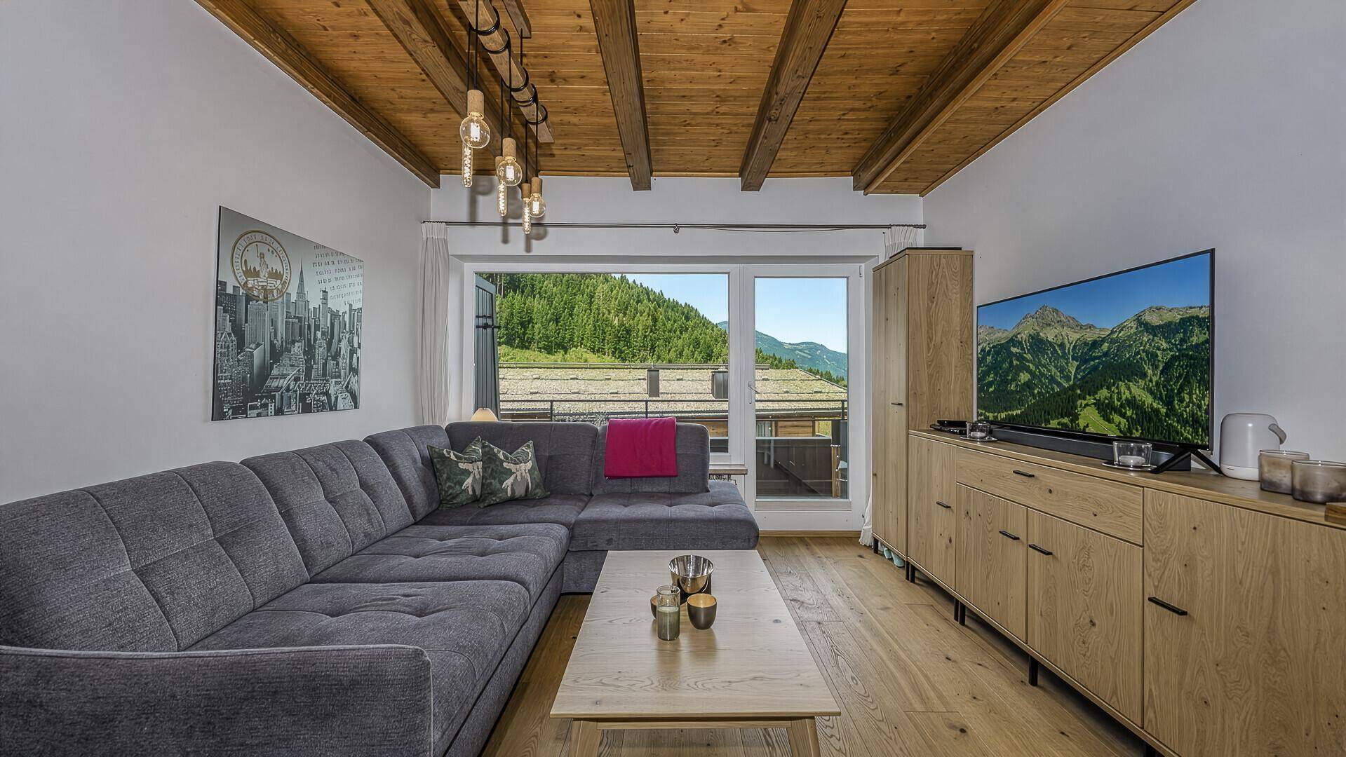 Gemütliches Wohnzimmer mit großem Sofa, Holzbalkendecke und weitem Blick auf die Berge.