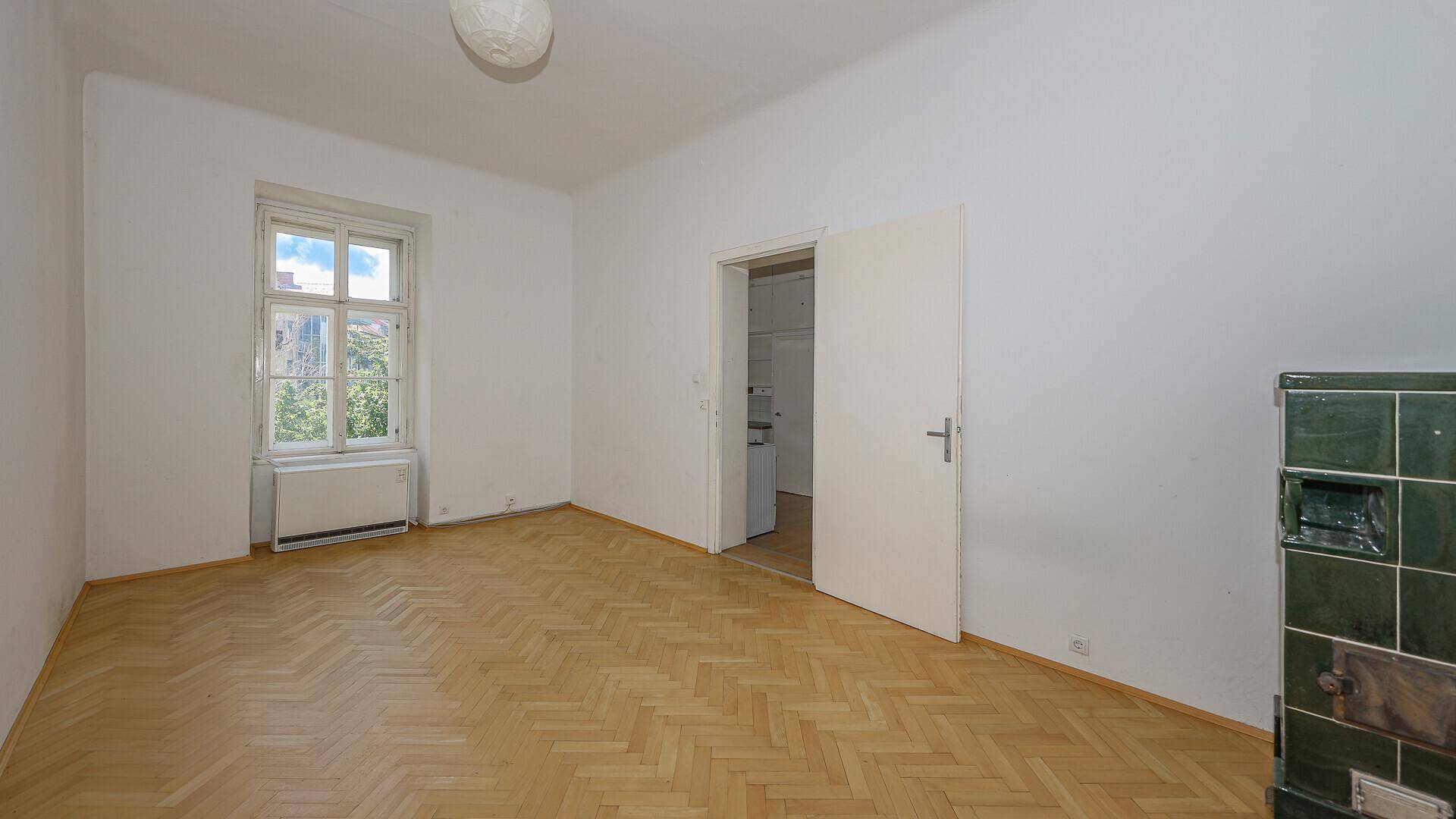 Kleineres Zimmer mit Fischgrätparkett, Fenster und einem traditionellen Kachelofen.