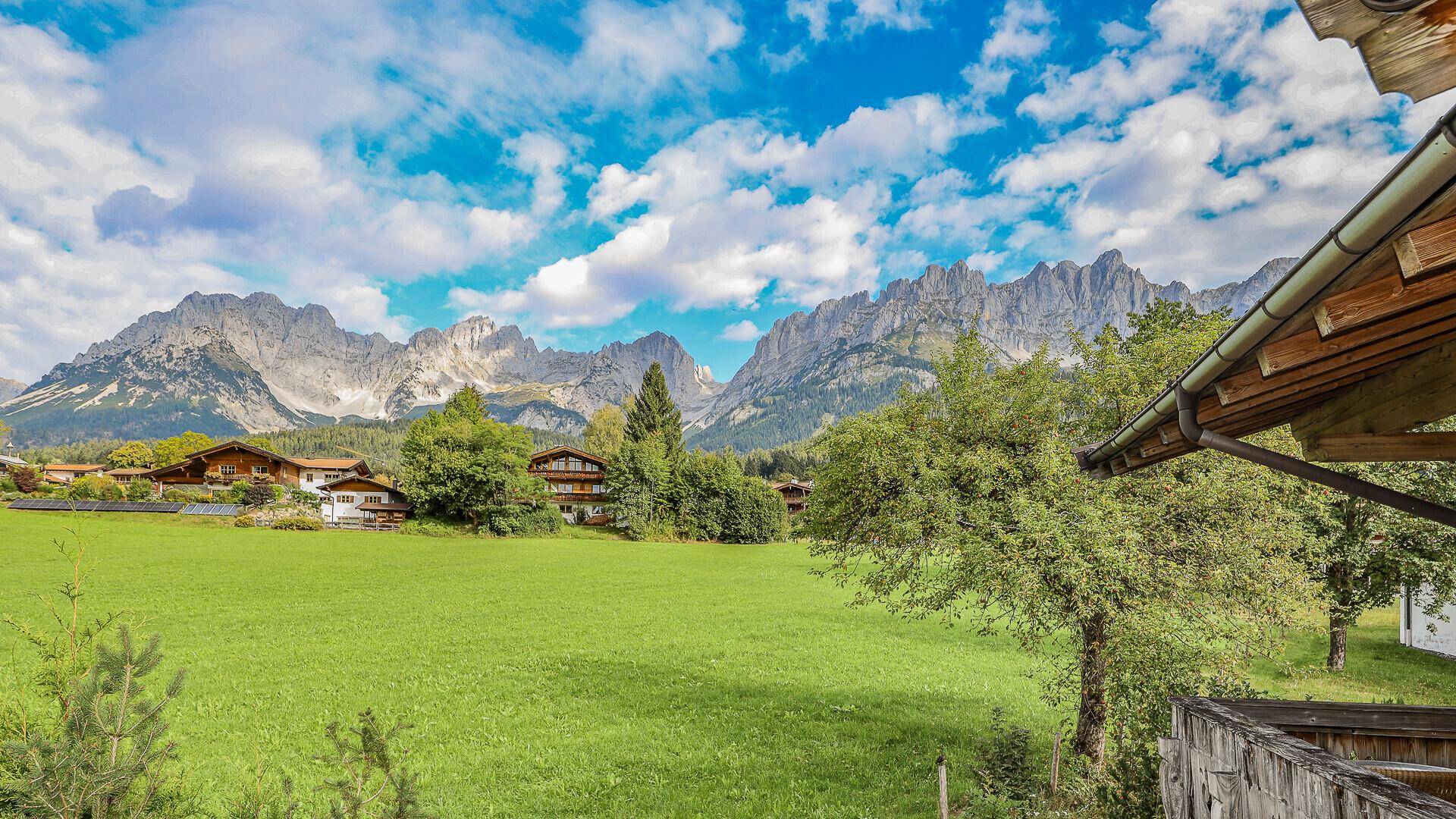 Weitläufiger Blick über grüne Wiesen und traditionelle Häuser mit majestätischer Bergkulisse im Hintergrund.