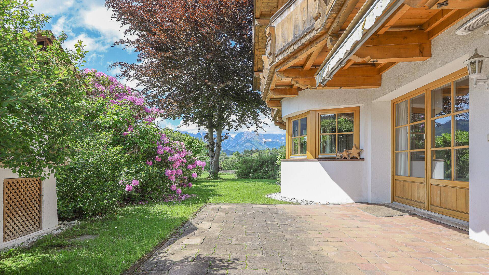 Gemütliche gepflasterte Terrasse am Haus, umgeben von blühenden Pflanzen mit Bergblick im Hintergrund.
