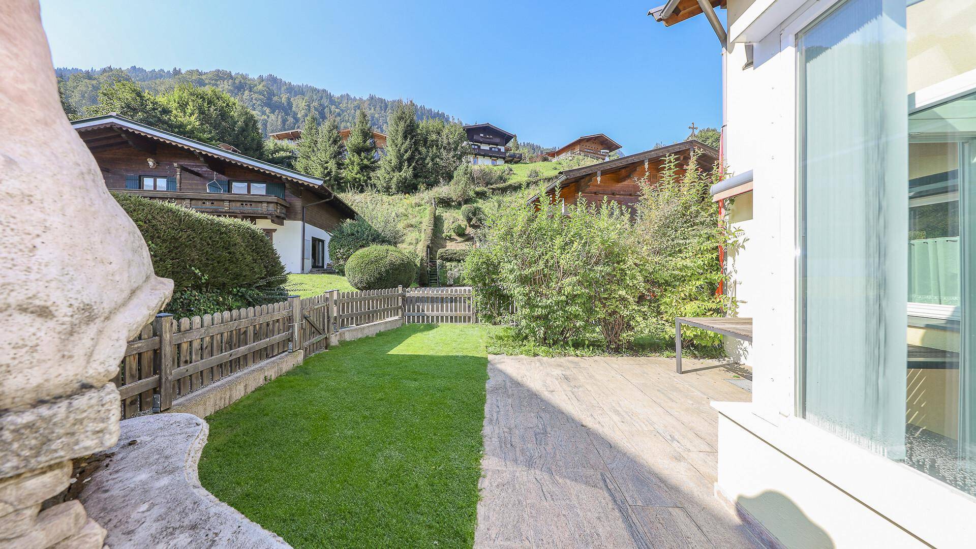 Gepflegter Gartenbereich mit Rasen, gepflasterter Terrasse und Blick auf die umliegenden Häuser.