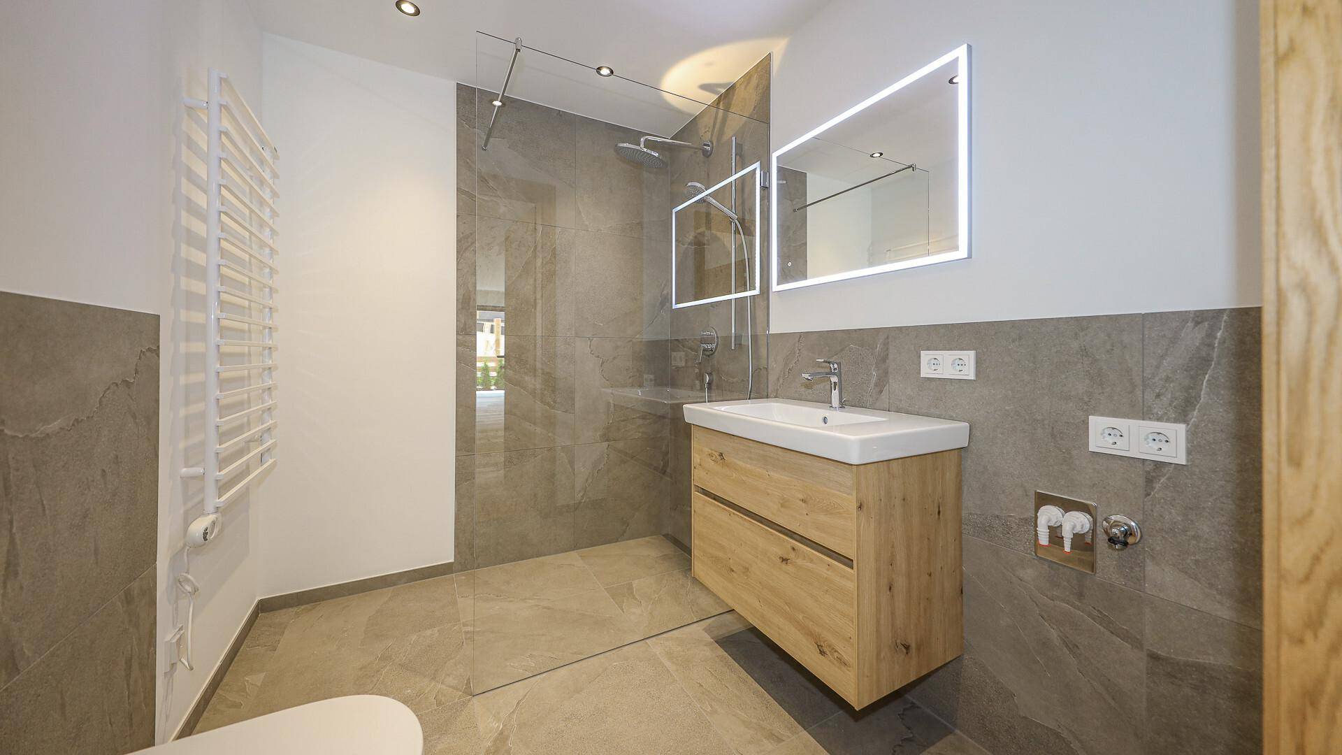 Modernes Badezimmer mit großformatigen Fliesen, Holzwaschtisch und beleuchtetem Spiegel.