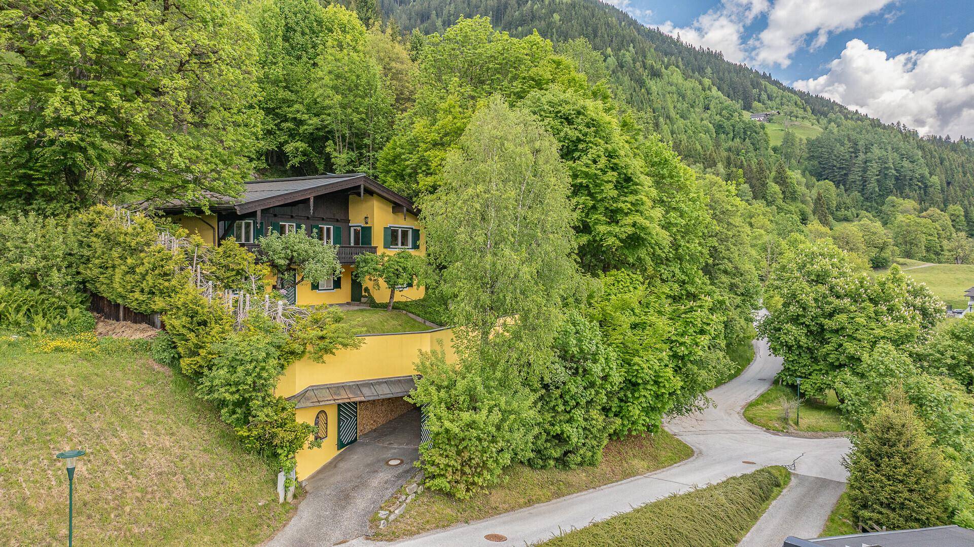 Haus in alpiner Umgebung mit Zufahrt, Carport und umgeben von viel Grün und Bergen.
