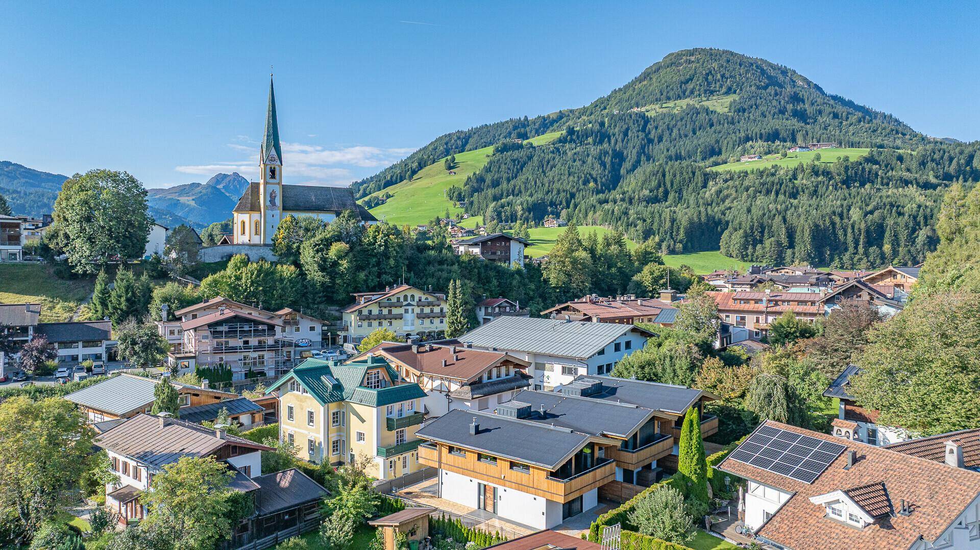Luftaufnahme eines charmanten Bergdorfes mit einer Kirche und umliegenden grünen Hügeln.
