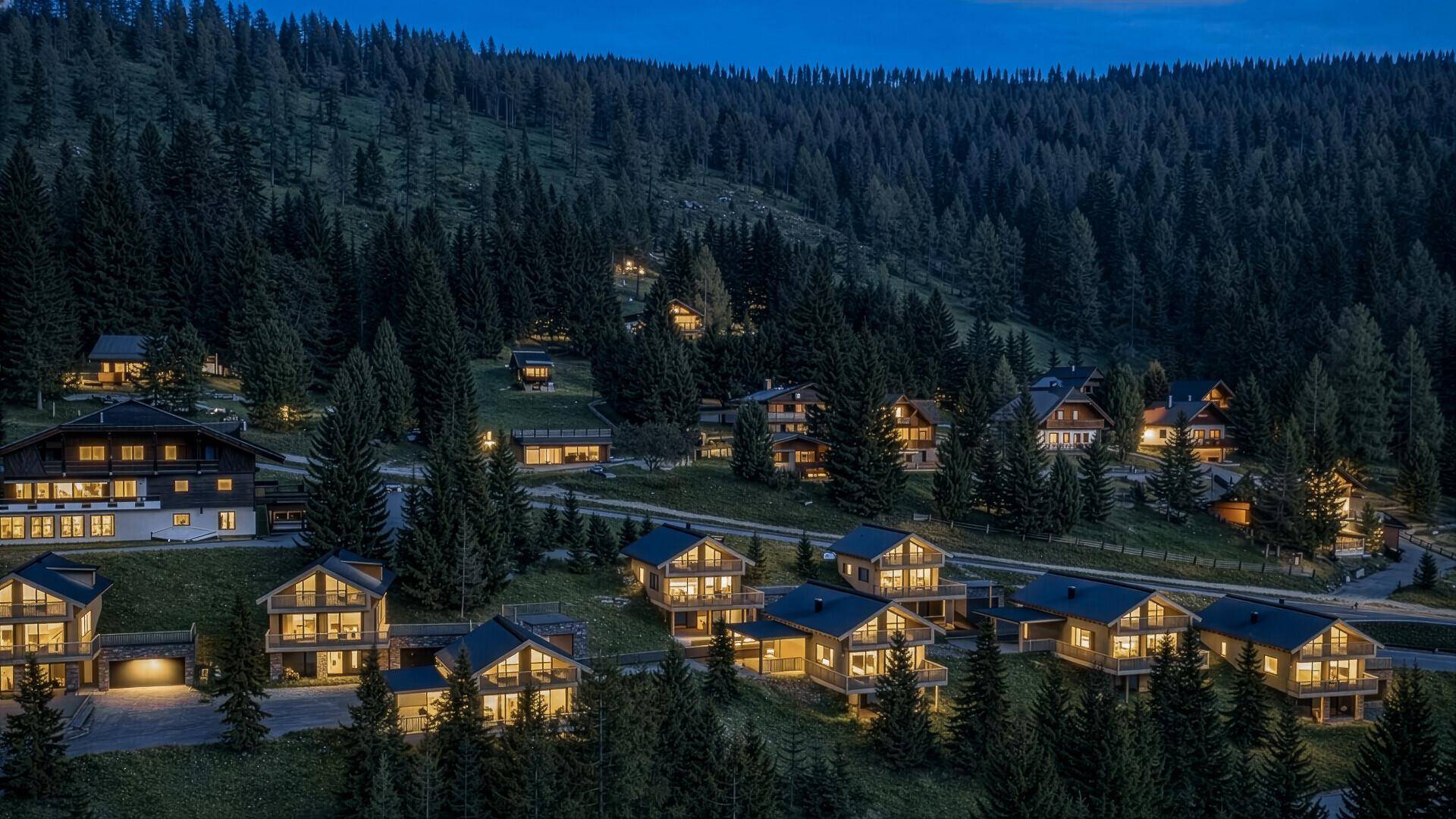 Abendliche Luftaufnahme einer Siedlung von beleuchteten Holzhäusern in einer bewaldeten Berglandschaft.