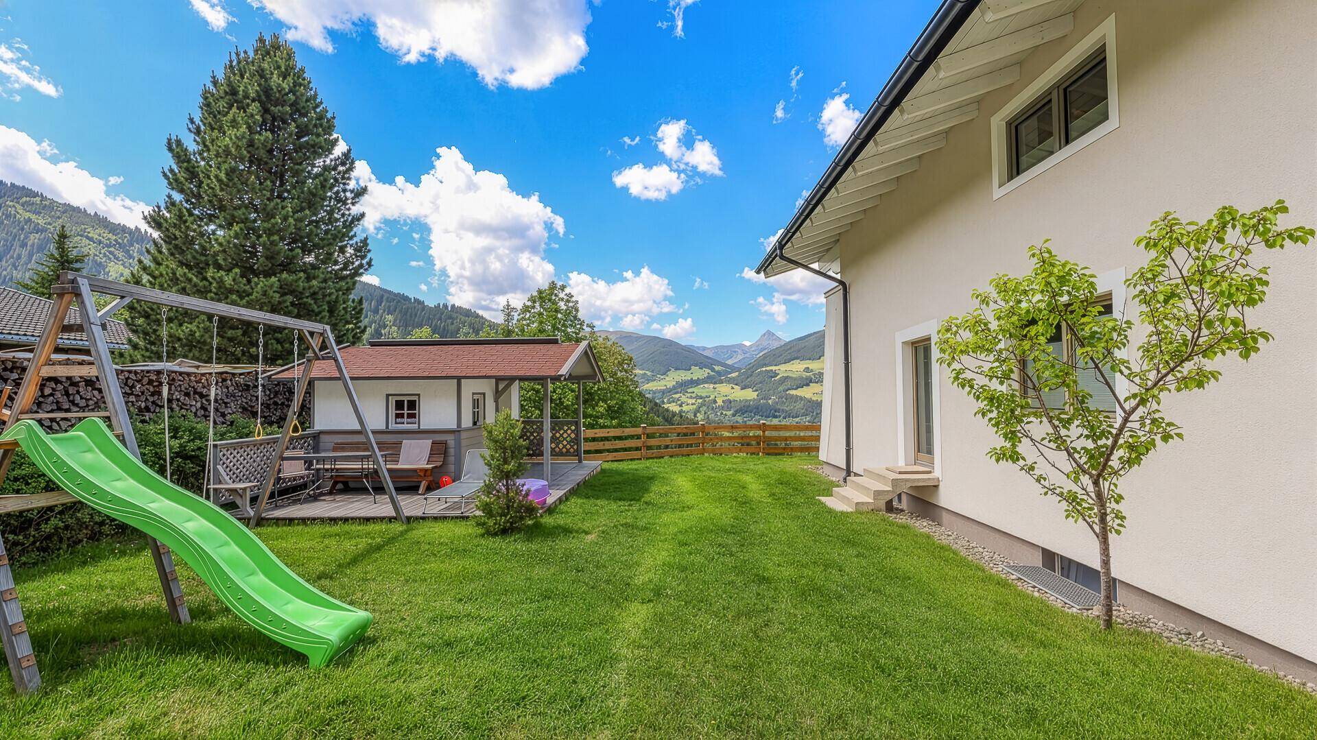 Gepflegter Garten mit Spielplatz, Rutsche und Schaukel, umgeben von einer malerischen Bergkulisse.