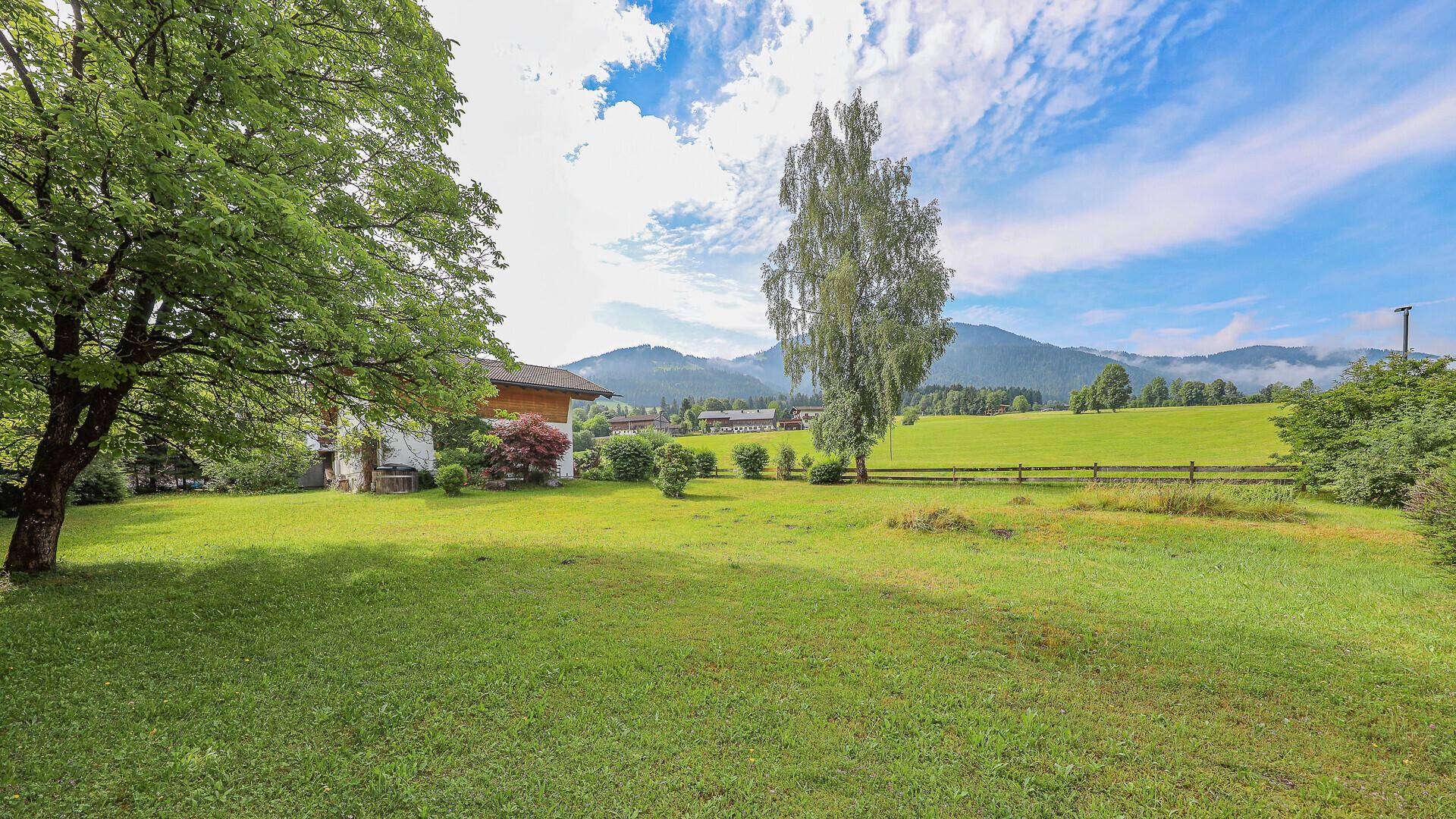 Weitläufiger Garten mit Rasenfläche und einzelnen Bäumen, umgeben von grüner Natur und Bergen.