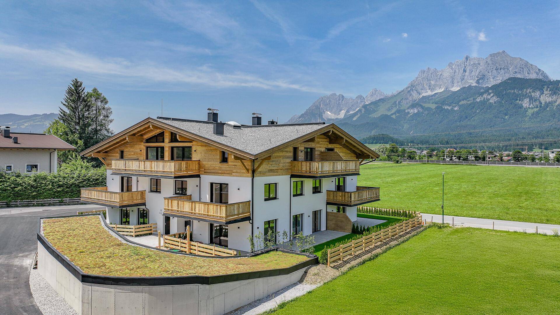 Neubau-Wohnhaus im alpinen Stil mit Holzbalkonen und Bergblick, umgeben von grünen Wiesen.